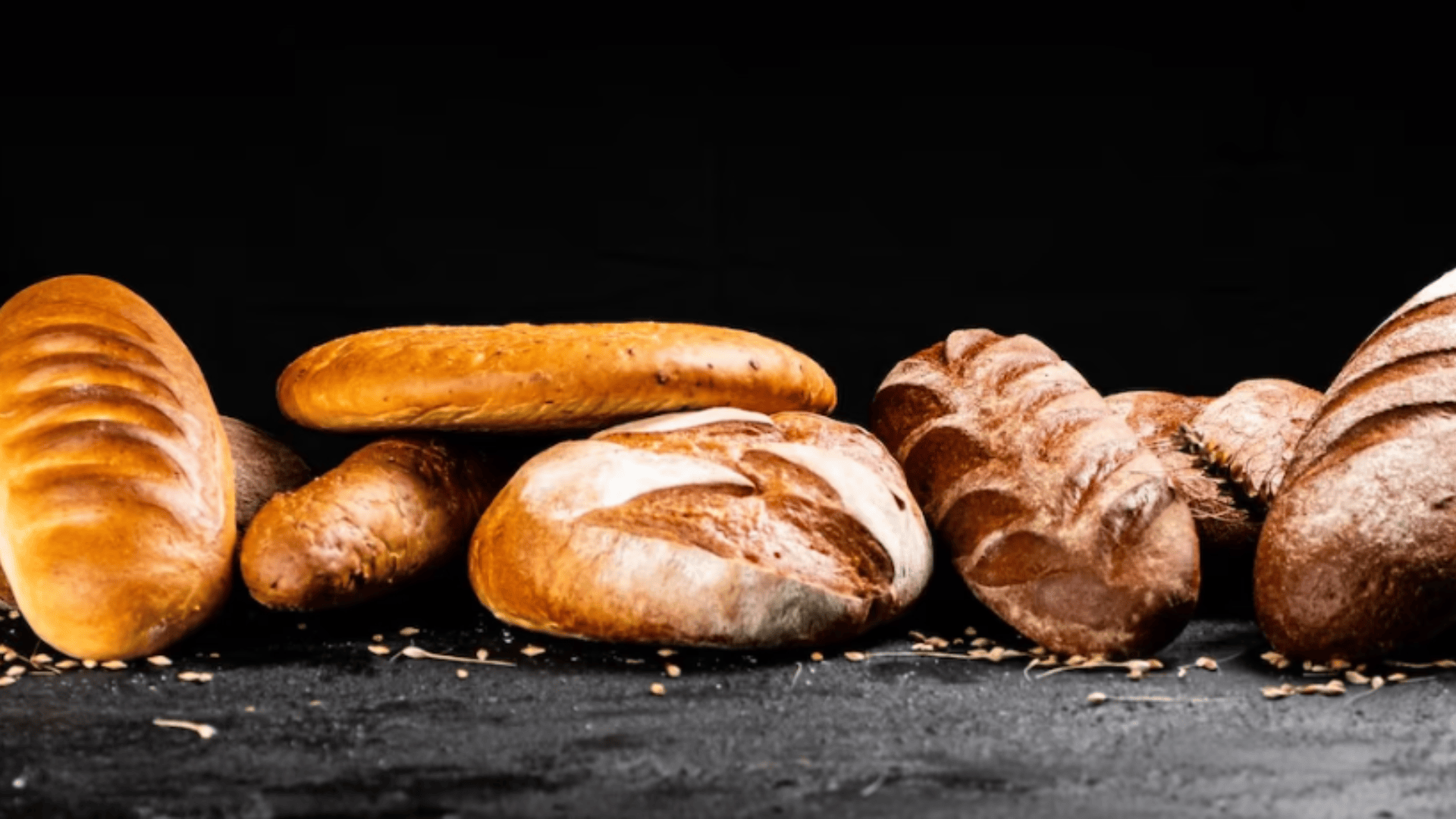 A variety of artisan bread loaves, including baguettes and round loaves, are arranged on a dark surface with scattered seeds, against a black background.