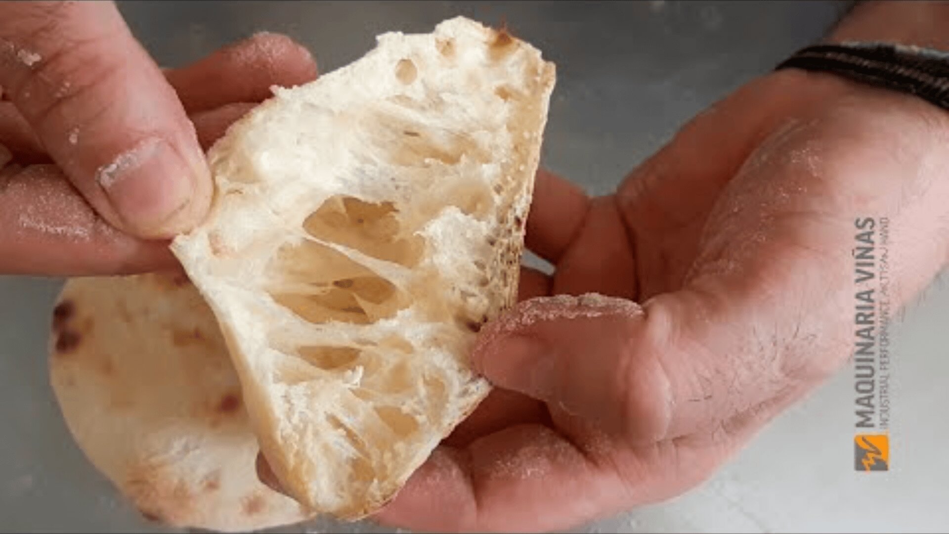 A close-up of two hands holding a piece of airy, rustic bread, showing its open, fluffy crumb and holes. The background is blurred and the Maquinaria Viñas logo is on the right side of the image.
