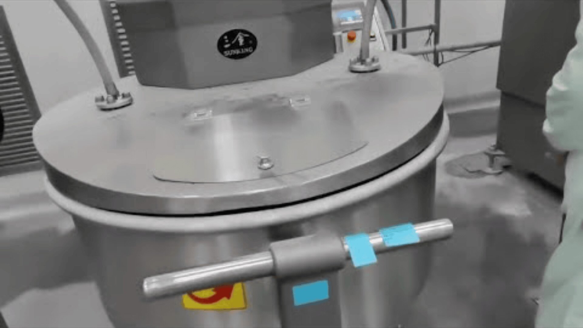 A large stainless steel industrial mixing machine with a closed lid is situated in a clean, bright facility. Control buttons and blue tape are visible on the front, with a person standing partially out of frame.