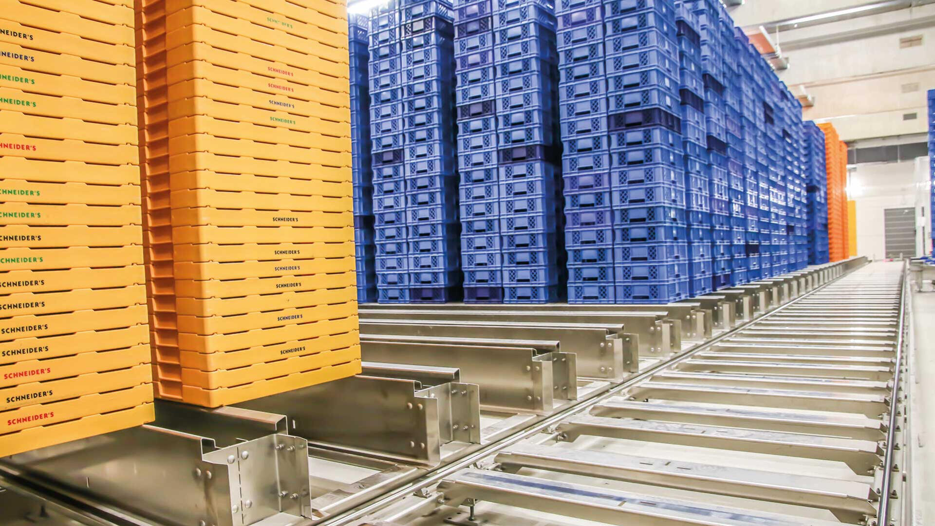 Stapel von orangefarbenen und blauen Kunststoffkisten sind in hohen Säulen auf einem Metallfördersystem in einem großen Lager oder einer Industrieanlage angeordnet.