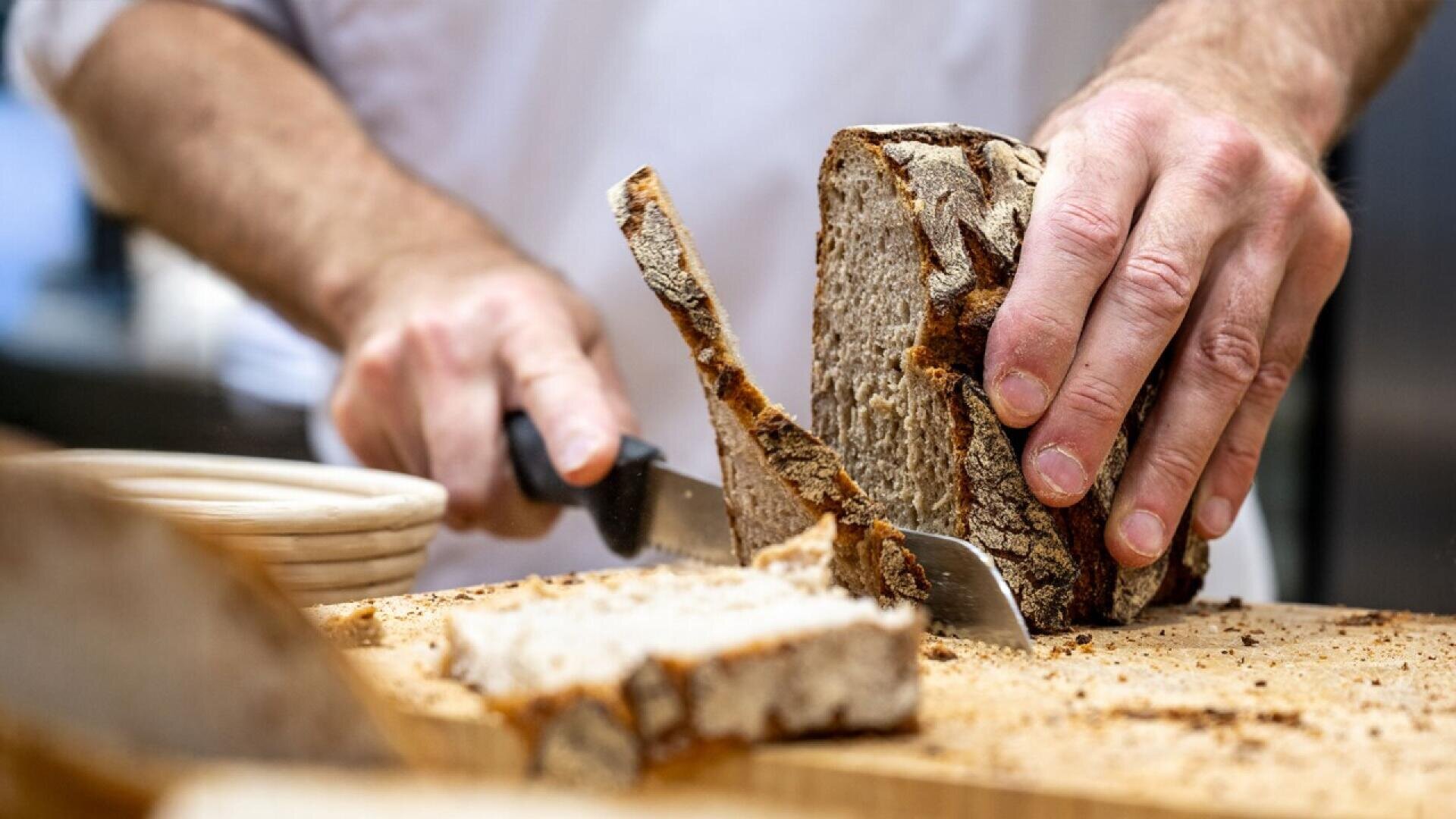 Close-up of a person slicing a loaf of rustic bread on a wooden cutting board with a serrated knife; hands and part of the torso are visible, with crumbs scattered on the board.