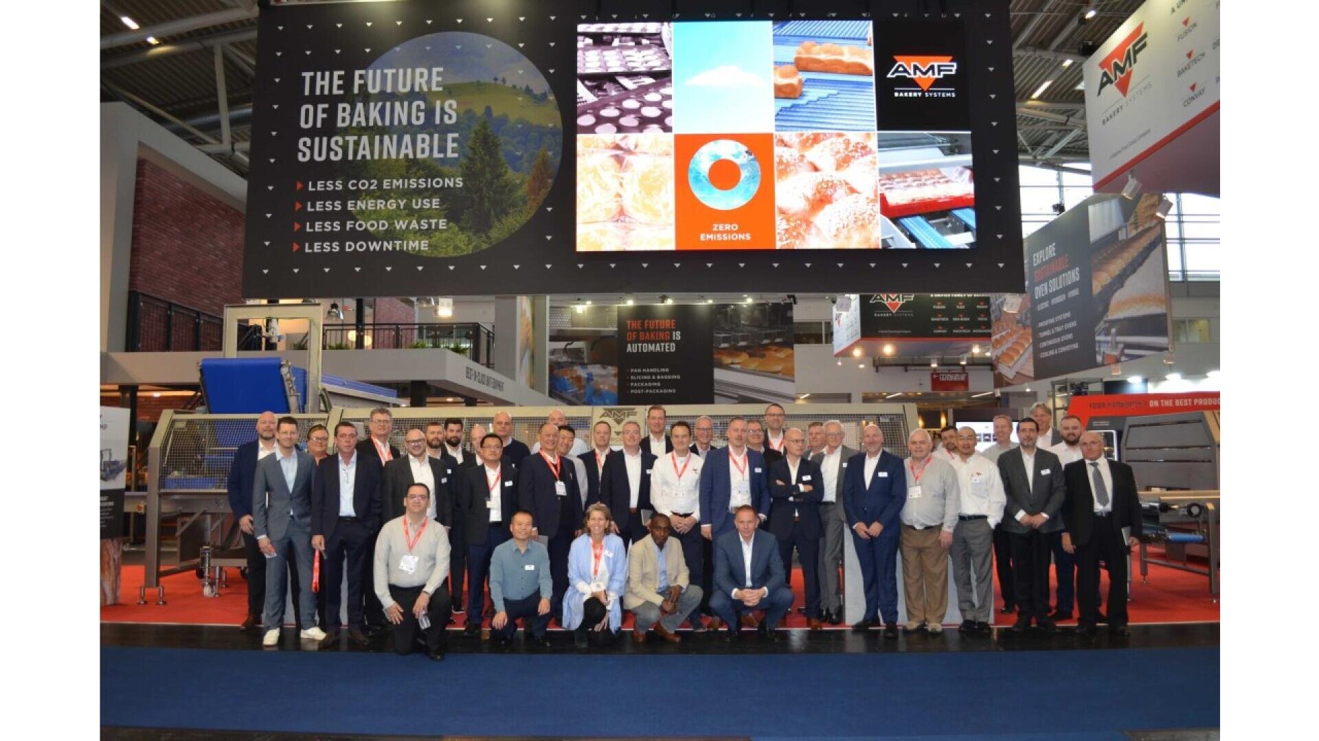 Eine Gruppe von Personen posiert für ein Foto auf einer Fachmesse vor großen Bildschirmen mit Botschaften zur Nachhaltigkeit beim Backen und Firmenlogos, umgeben von Messeständen und Bäckereimaschinen.