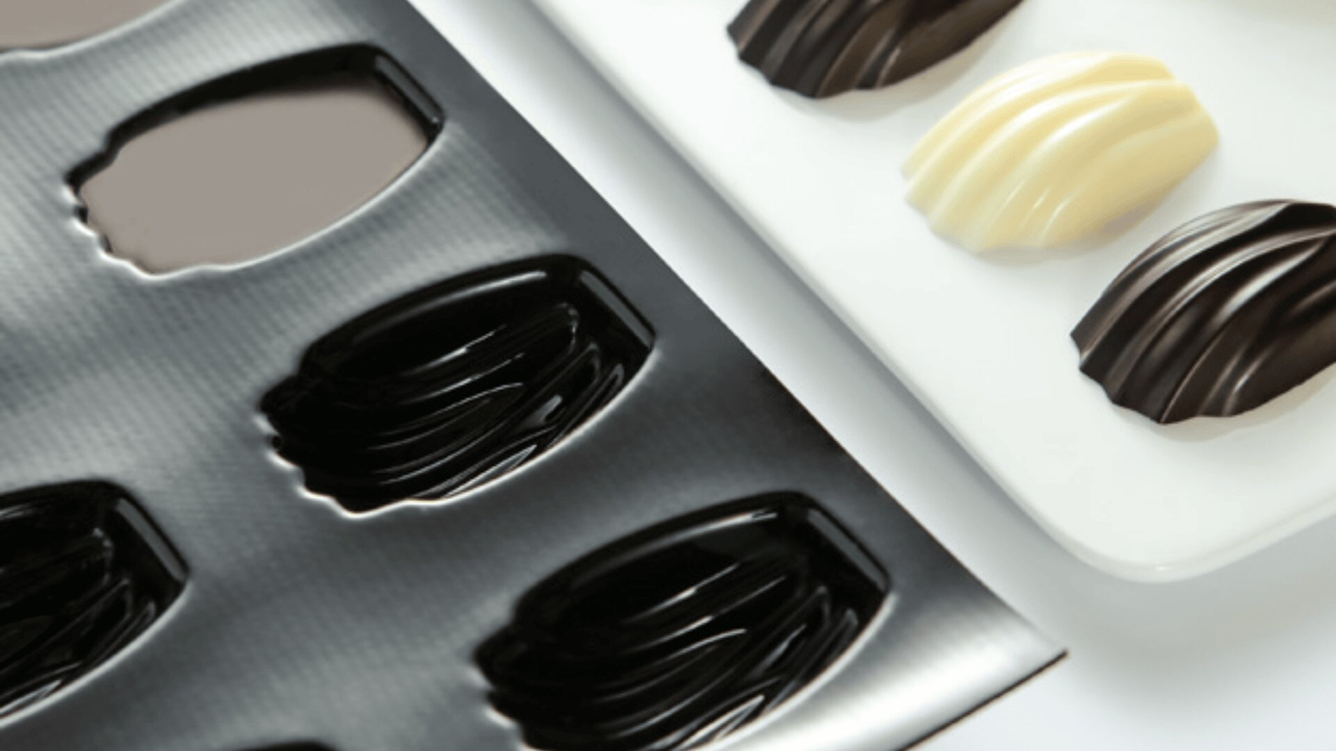 A close-up of a black plastic chocolate mold tray next to a white plate holding three shell-shaped chocolates—two dark and one white.