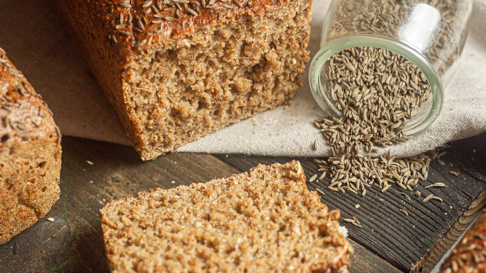 A loaf of whole grain bread topped with caraway seeds sits next to a slice of bread. A small glass jar on its side spills caraway seeds onto a rustic wooden surface.