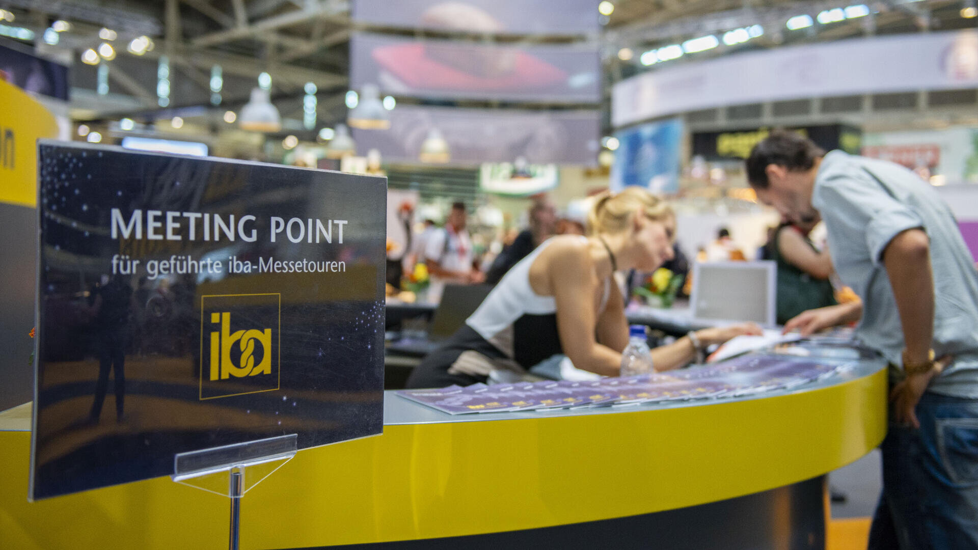 Ein Schild mit der Aufschrift MEETING POINT für geführte iba-Messetouren steht vor einem gebogenen Tresen, an dem in einer belebten Messehalle Gespräche geführt und Dokumente eingesehen werden.