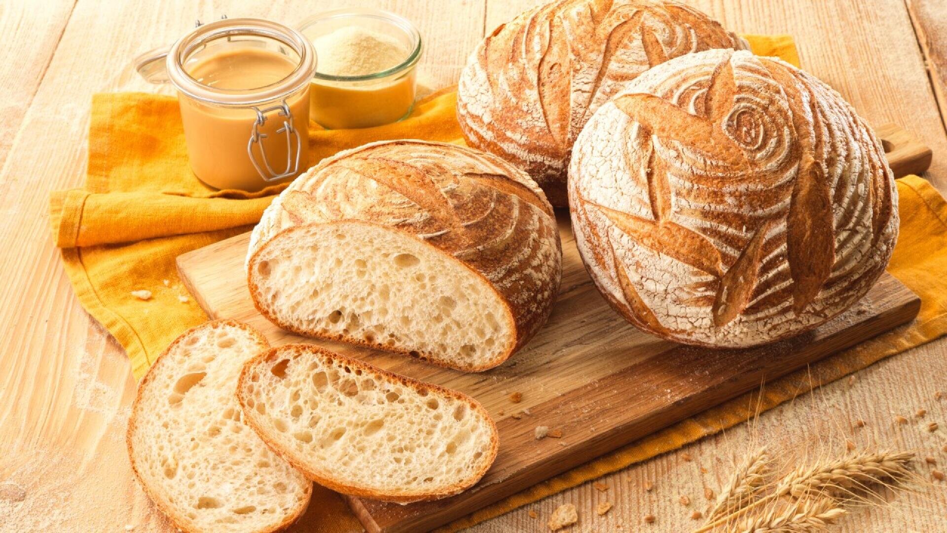 Two round artisan sourdough loaves and several slices rest on a wooden cutting board. Jars of flour and spread sit nearby on a yellow cloth, with wheat stalks on a rustic wooden table.