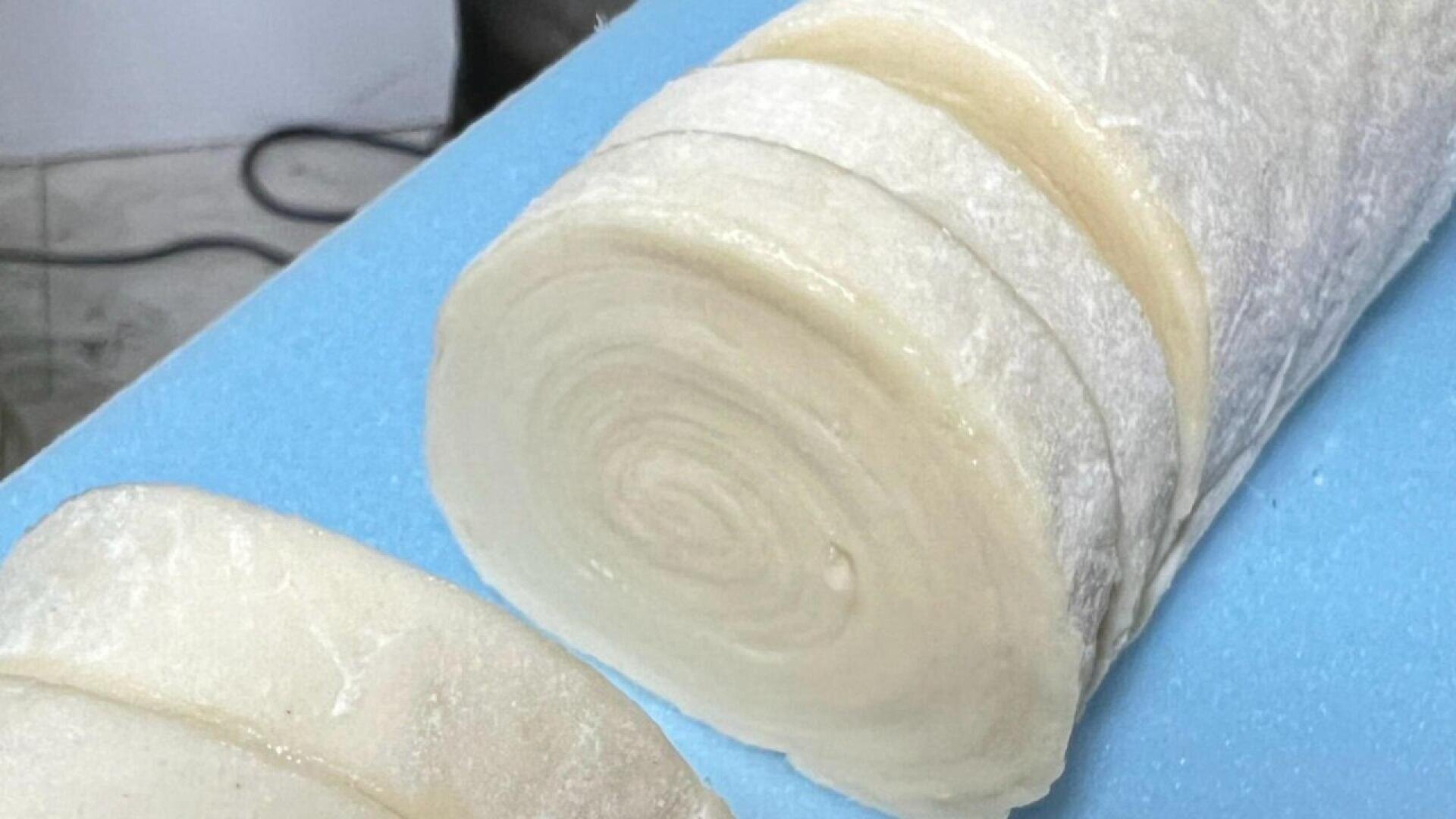 Eine Nahaufnahme des gerollten Teigs, der in dicke, gleichmäßige Stücke geschnitten und auf einem blauen Schneidebrett präsentiert wird. Der Teig hat ein spiralförmiges Muster und ist an der Oberfläche mit Mehl bestäubt.