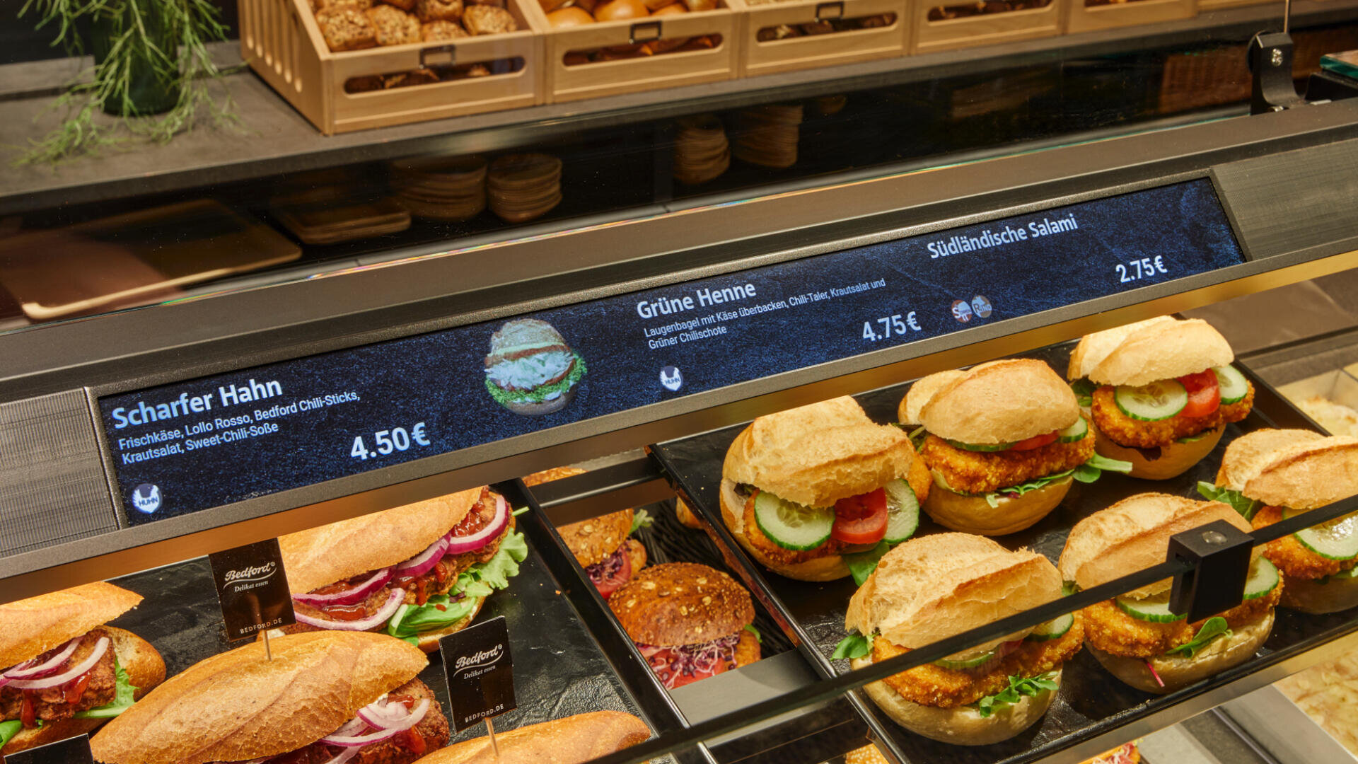 Eine gläserne Bäckereivitrine zeigt Sandwiches mit Brötchen, Salat, Tomate und Fleisch. Digitale Etiketten darüber geben Namen und Preise an: Scharfer Hahn, Grüne Henne und Südtiroler Salami.