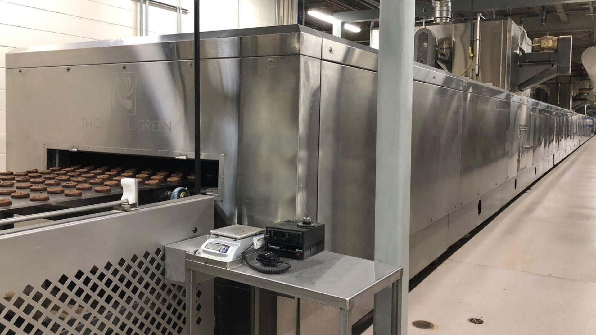 A large industrial baking oven with conveyor belts moves rows of cookies through the machine in a clean, modern factory setting. Nearby, a metal table holds a scale and electronic equipment.