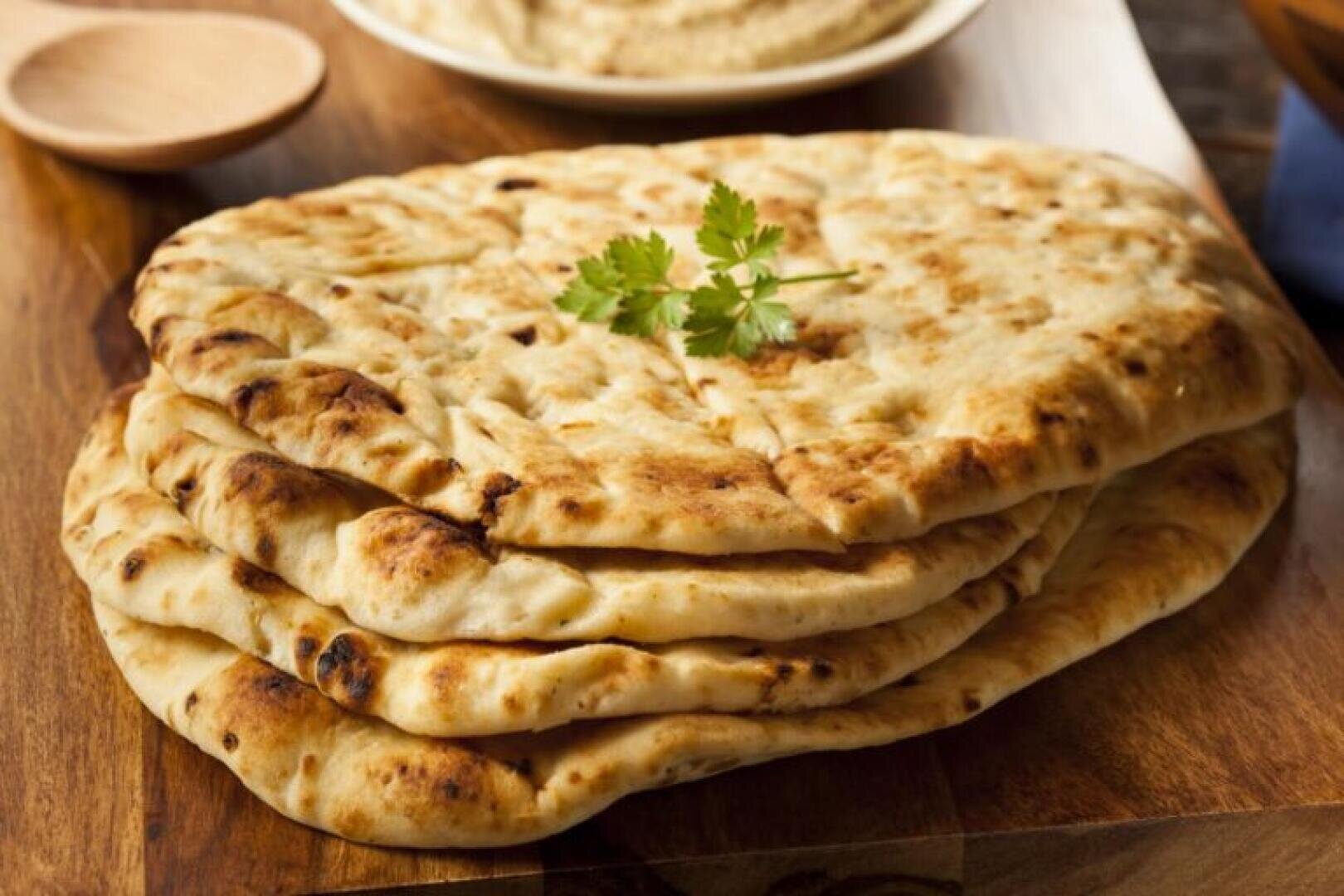 A stack of naan bread on a wooden board, topped with a sprig of cilantro, with a wooden spoon and a dish of hummus in the background.