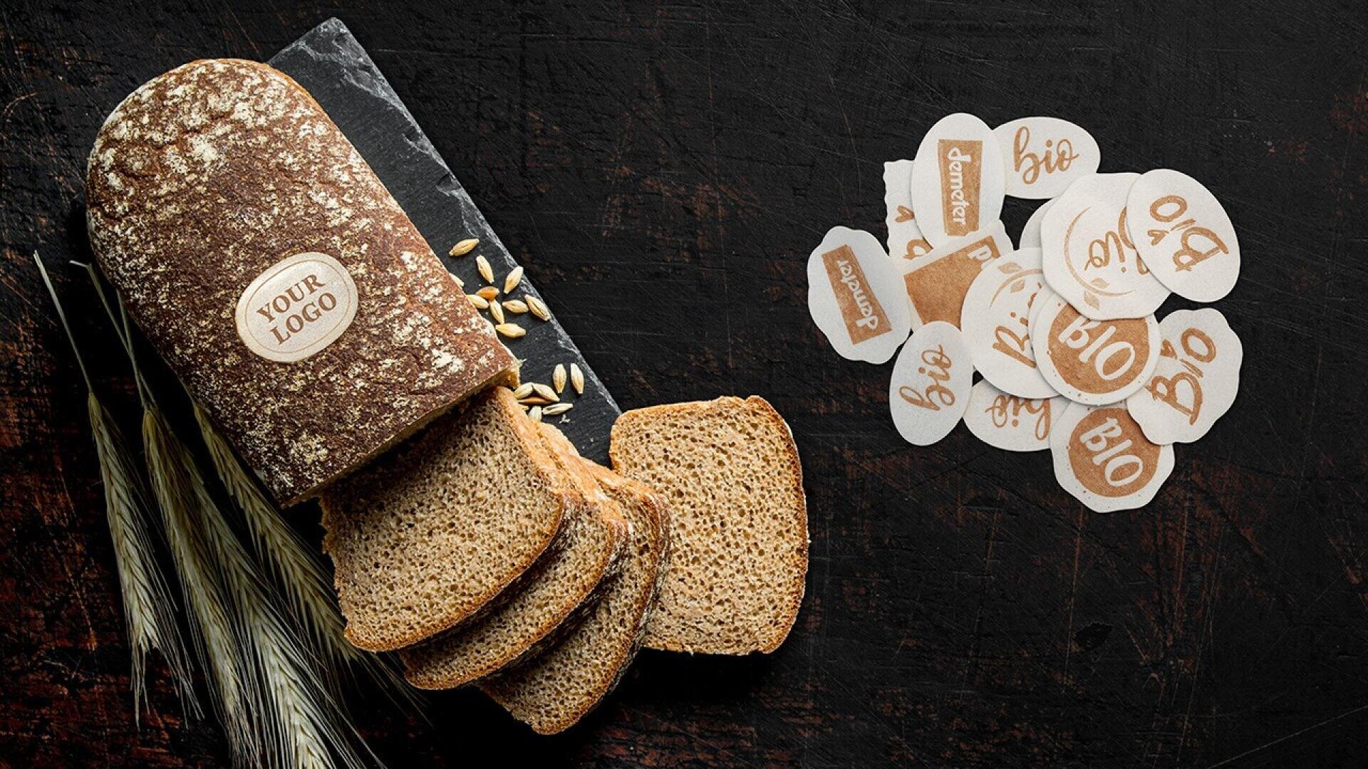 A loaf of sliced whole wheat bread on a dark surface, with wheat stalks and grains nearby. Several round and oval labels reading bio and demeter are spread out to the right of the bread.