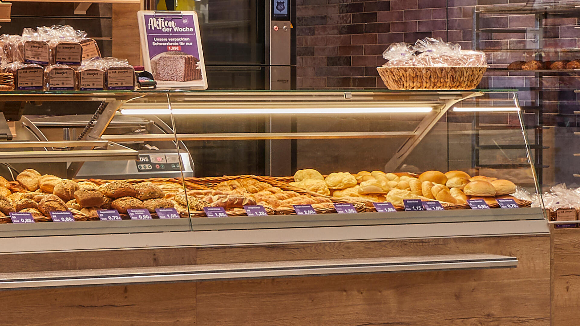 Eine Bäckereivitrine mit verschiedenen Brotsorten und Brötchen, darunter Baguette, Saatenbrot und Brötchen, mit sichtbaren Preisschildern auf dem Glas und verpackten Broten in den Regalen darüber.