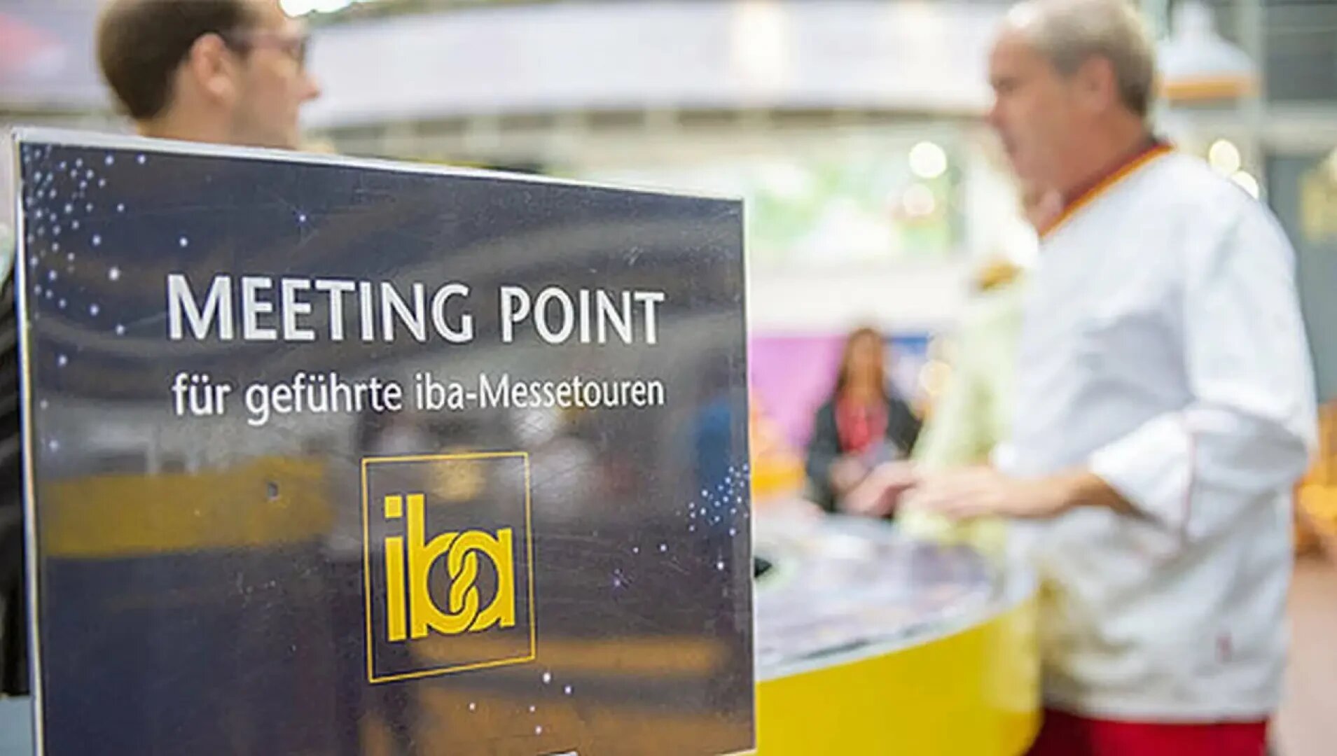 Ein Schild mit der Aufschrift MEETING POINT für geführte iba-Messetouren steht im Mittelpunkt, während sich zwei Personen, eine davon in einer Kochuniform, im unscharfen Hintergrund bei einer Indoor-Veranstaltung unterhalten.