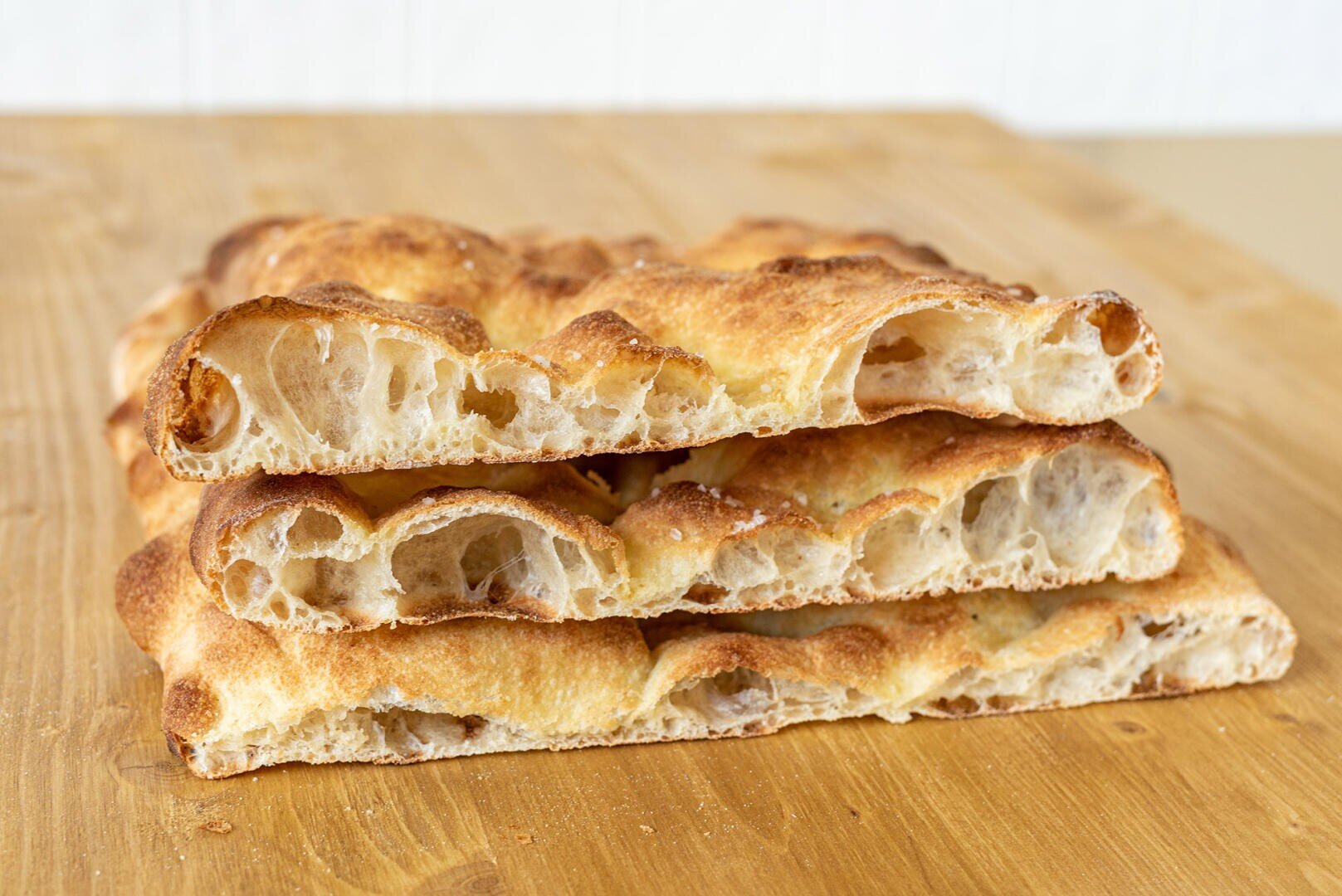 Three pieces of airy, golden-brown focaccia bread are stacked on a wooden surface, showing a crisp crust and open crumb with large holes inside.