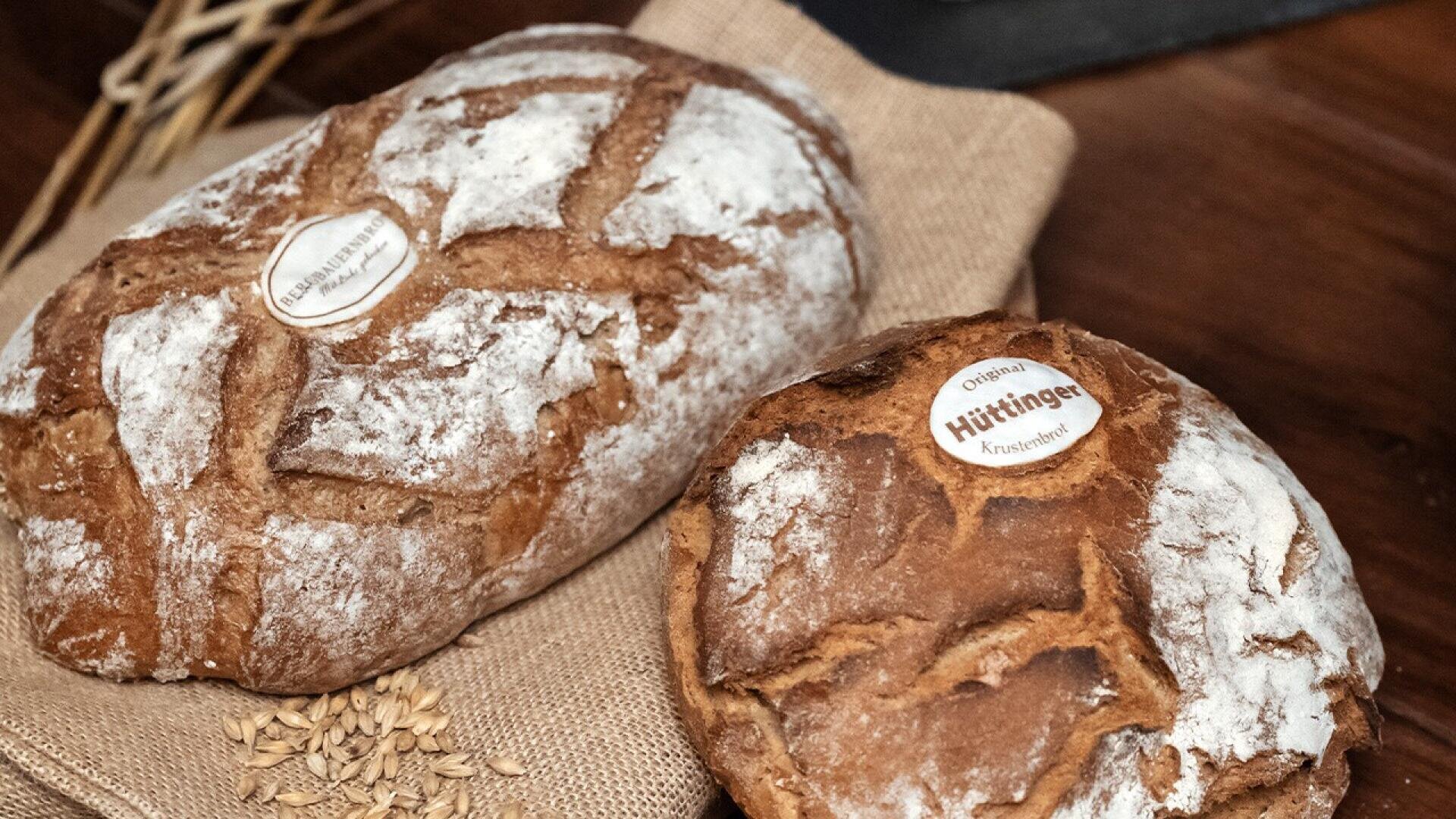 Two round loaves of rustic bread with cracked crusts and dusted with flour lie on a burlap cloth. Each loaf has a small round label on top. Some wheat grains are scattered nearby.