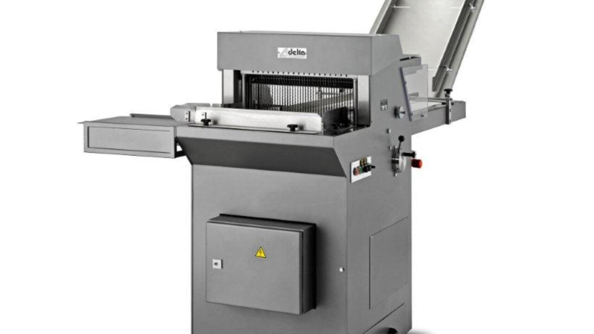 A large, industrial bread slicing machine with a stainless steel finish, open top lid, control buttons on the side, and a tray for collecting sliced bread.