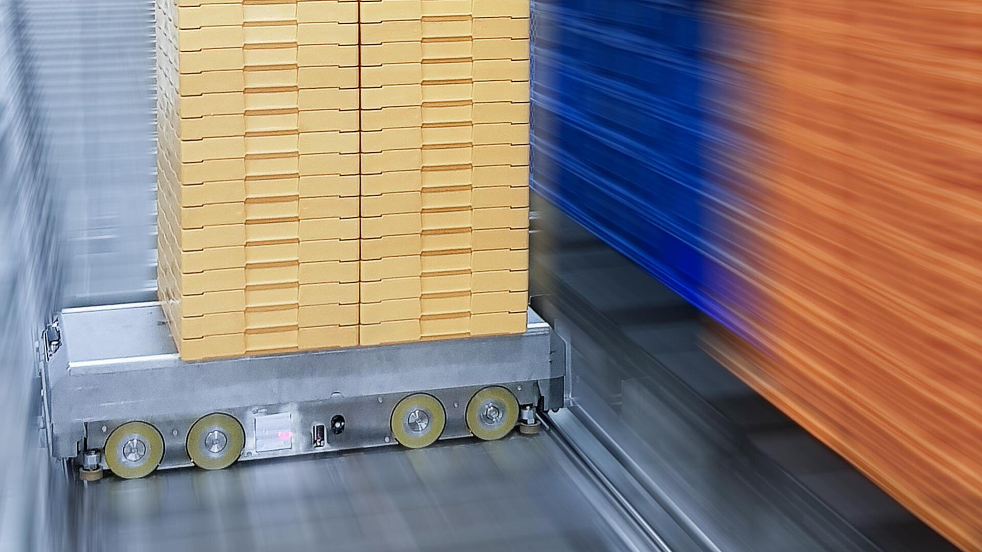 Ein Roboterwagen transportiert einen großen Stapel gelber Kartons in einem Lagerhaus, wobei die Bewegungsunschärfe die Geschwindigkeit und Effizienz betont; im Hintergrund sind blaue und orangefarbene Stapel zu sehen.