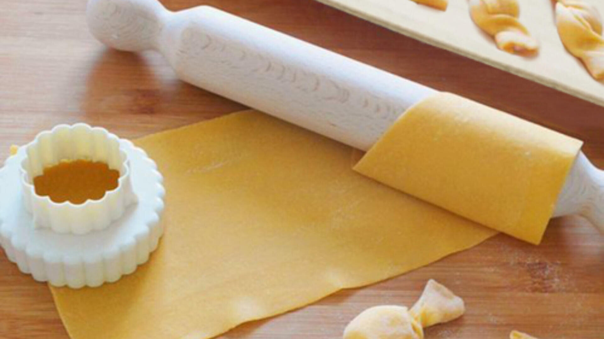 A wooden rolling pin rests on a sheet of pasta dough beside a round, scalloped-edged pasta cutter on a wooden surface. Pieces of shaped pasta are visible nearby.