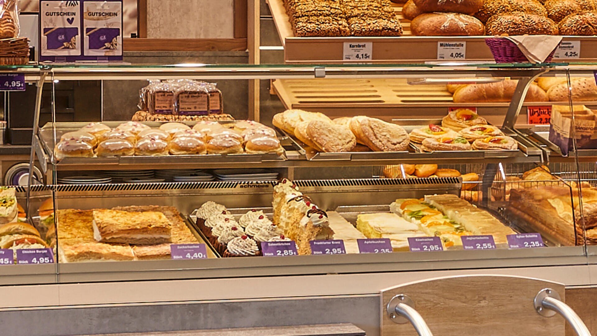 Eine Bäckereivitrine, gefüllt mit verschiedenen Gebäcken, Kuchen und Broten, mit sichtbaren Preisschildern. Hinter dem Glas sind Brote, Sandwiches, Sahnetorten und andere Backwaren ordentlich auf Tabletts und Regalen angeordnet.