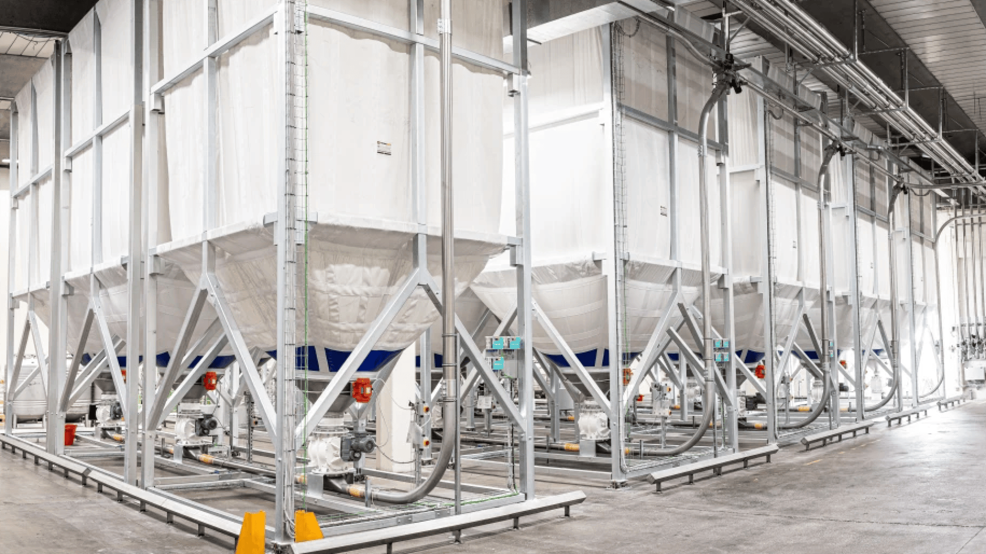 Large white industrial storage silos with metal frames inside a spacious, well-lit facility. Pipes and machinery are connected to the silos, and the floor appears clean and organized.