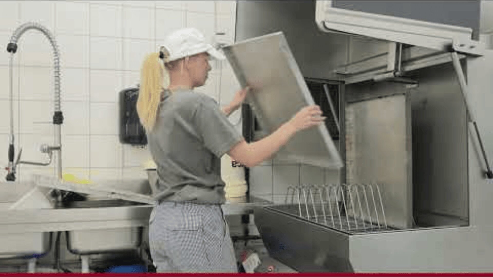 A person wearing a white cap and gray shirt loads a large tray into a commercial dishwasher in an industrial kitchen with tiled walls and a steel sink.