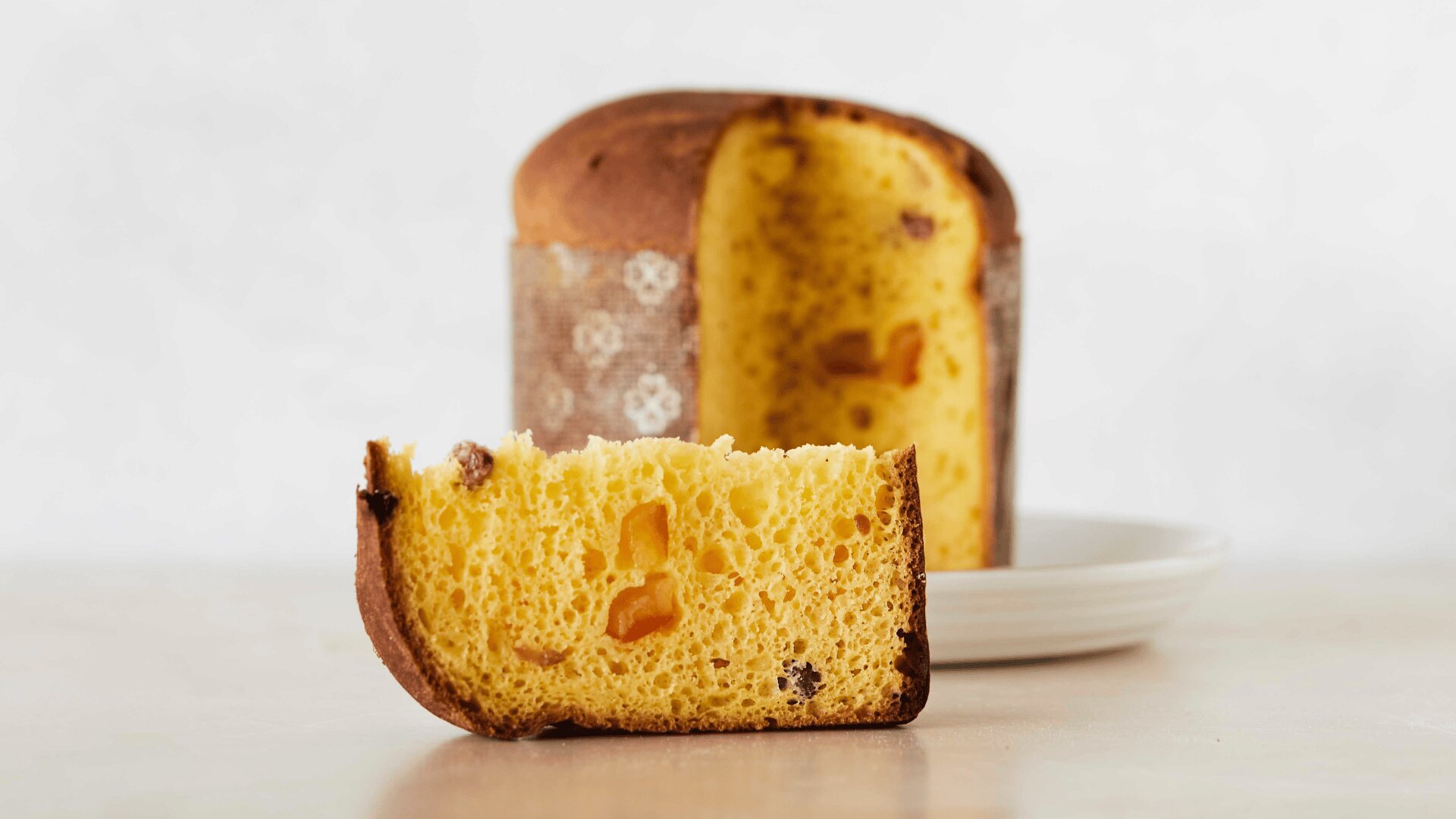 A sliced panettone, an Italian sweet bread with visible dried fruit pieces, sits on a plate against a plain, light background. One piece is placed in front of the rest of the loaf.