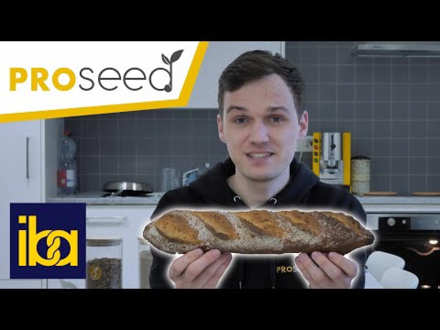 A person in a kitchen holds a loaf of bread. The image has the PROseed logo in the top left and the iba logo in the bottom left. The background features kitchen appliances and containers.