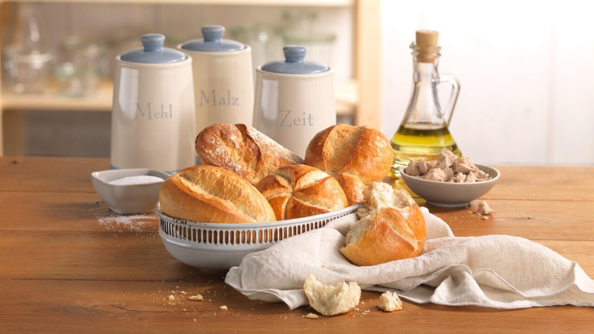 A bowl of freshly baked bread rolls sits on a wooden table with a beige cloth. Nearby are a bottle of oil, a bowl of yeast, a dish of salt, and three ceramic jars labeled Mehl, Malz, and Zeit.