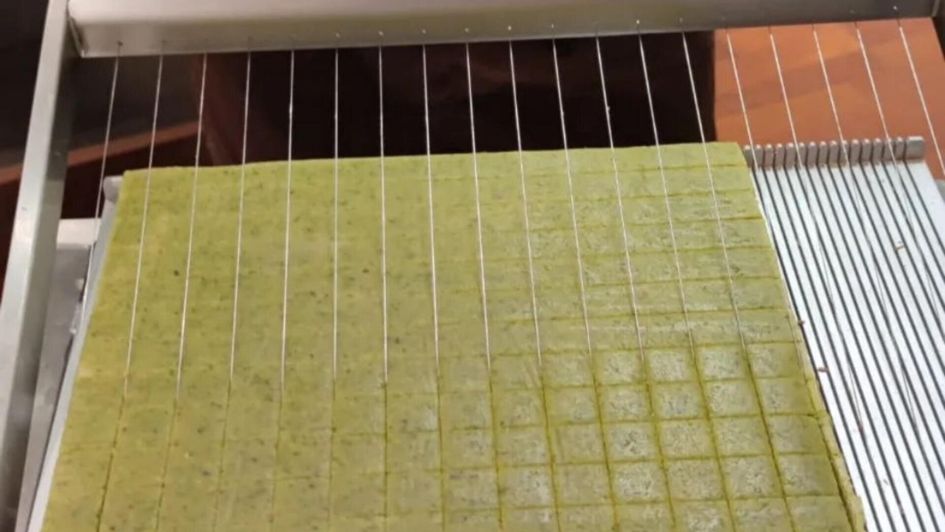 Eine Drahtschneidemaschine schneidet ein großes, flaches Blatt grünen Teig in gleichmäßig große Quadrate, wodurch ein Gittermuster entsteht. Die Schneidemaschine hat mehrere parallele Drähte, die über einen Metallrahmen gespannt sind.