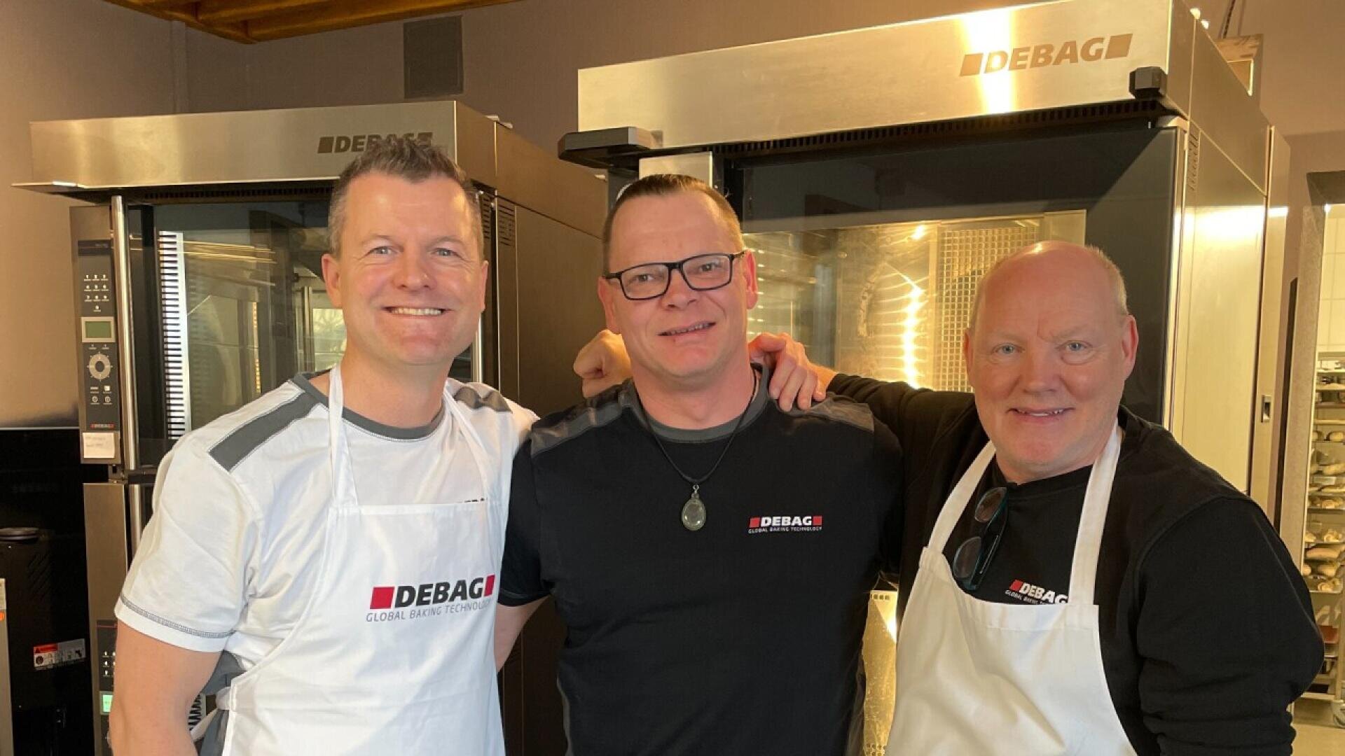 Three smiling men stand together in a bakery kitchen, two wearing white aprons and all wearing DEBAG branded shirts, with large bakery ovens in the background.