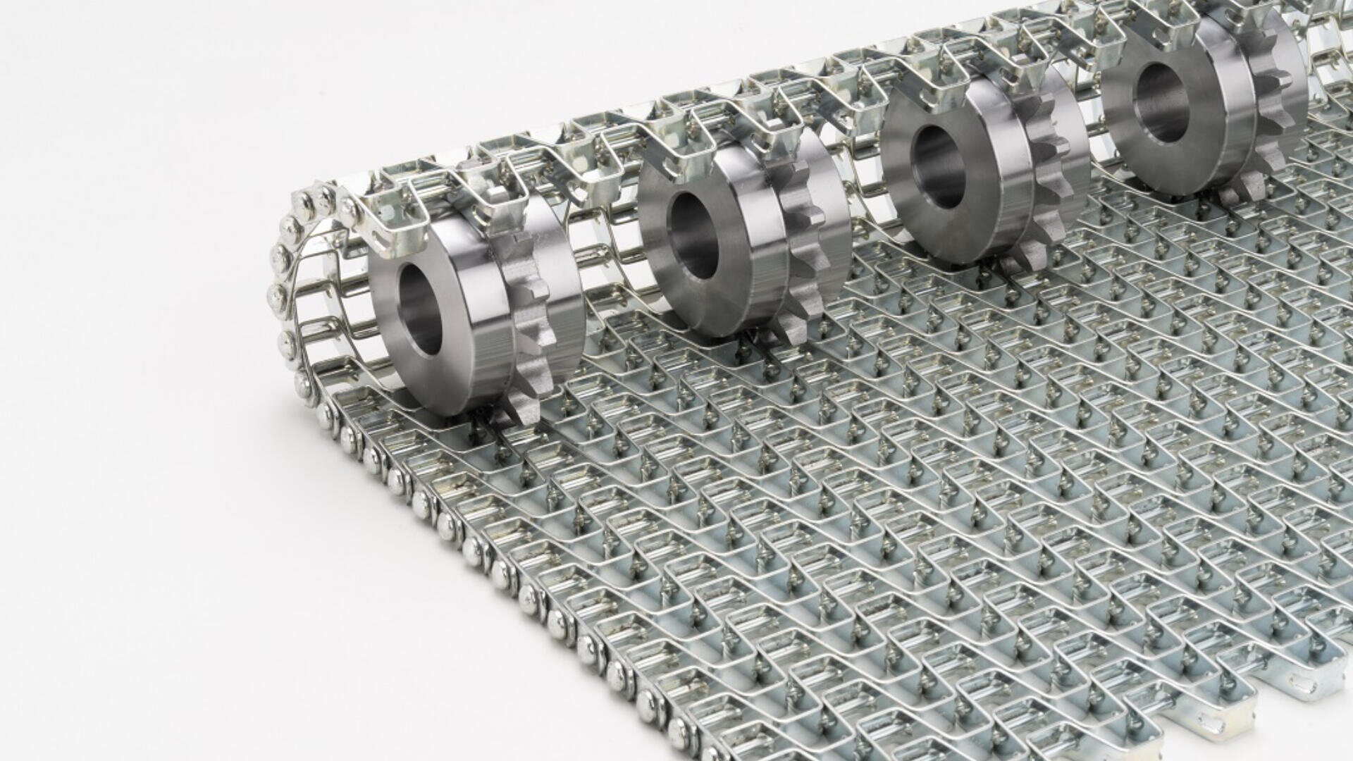 Close-up of a metal conveyor belt with interlocking wire mesh links and four evenly spaced metal sprockets on top, designed for industrial use. The background is plain white.