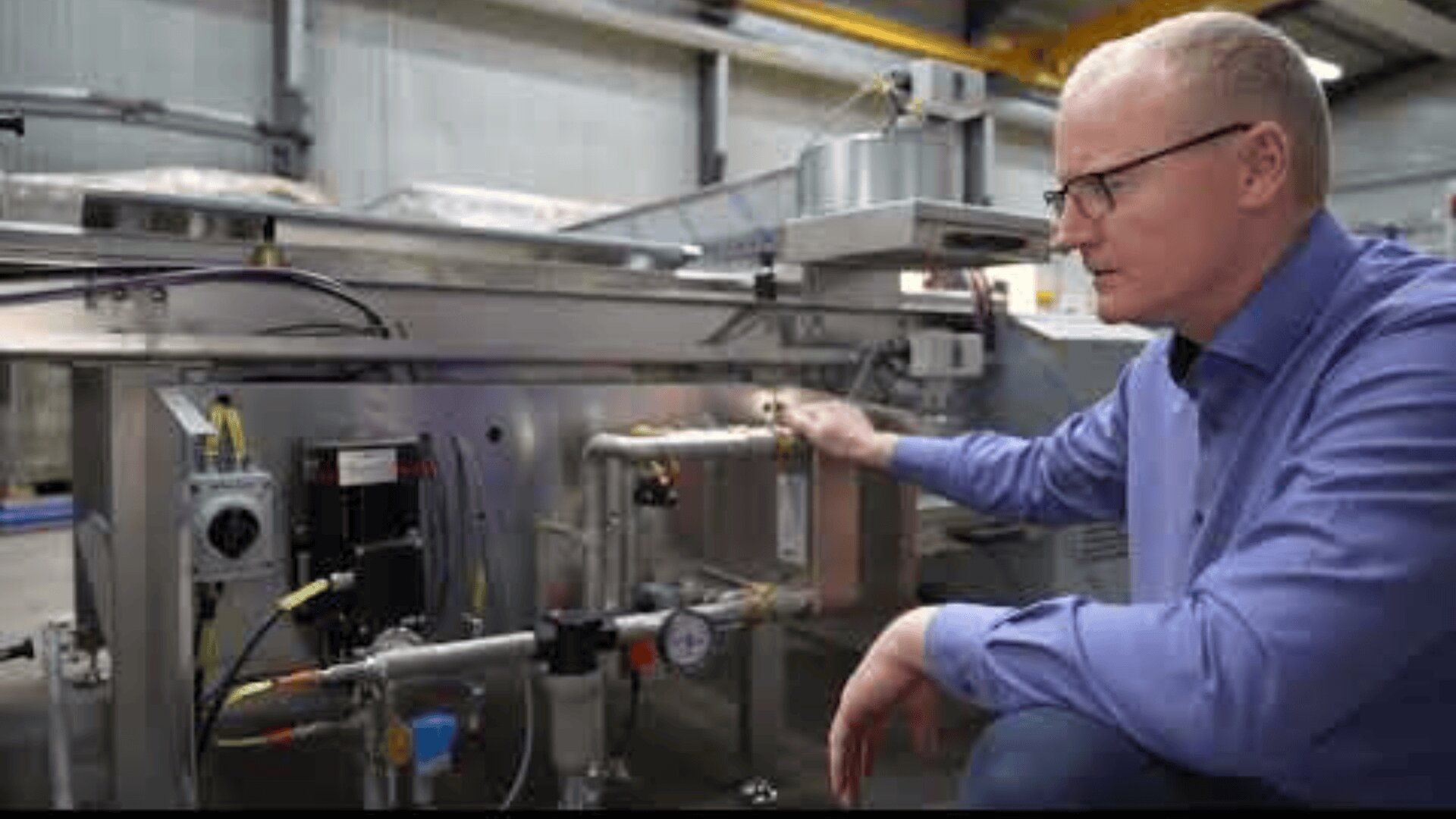 Ein Mann mit Brille und blauem Hemd hockt in einer Fabrik neben Industriemaschinen und überprüft Rohre und Messgeräte, die an den Metallgeräten angebracht sind.