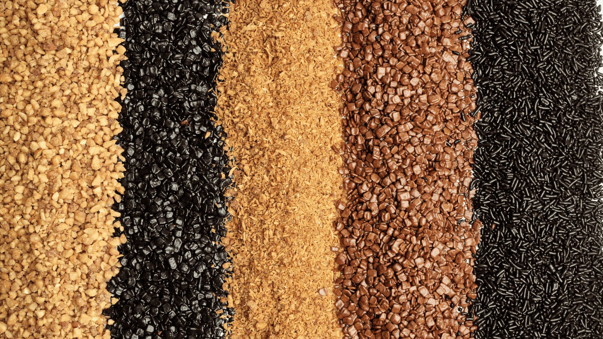 Rows of various textured plastic granules and pellets in different colors, including tan, black, light brown, dark brown, and dark gray, arranged vertically side by side.
