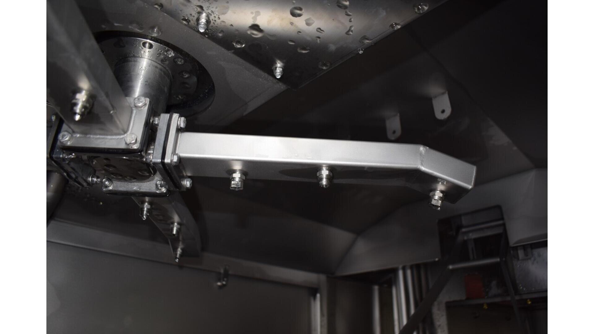 Close-up view of a metallic industrial mixing or processing machine part with bolts and a rotating arm, inside a polished metal chamber with water droplets and structural components visible.