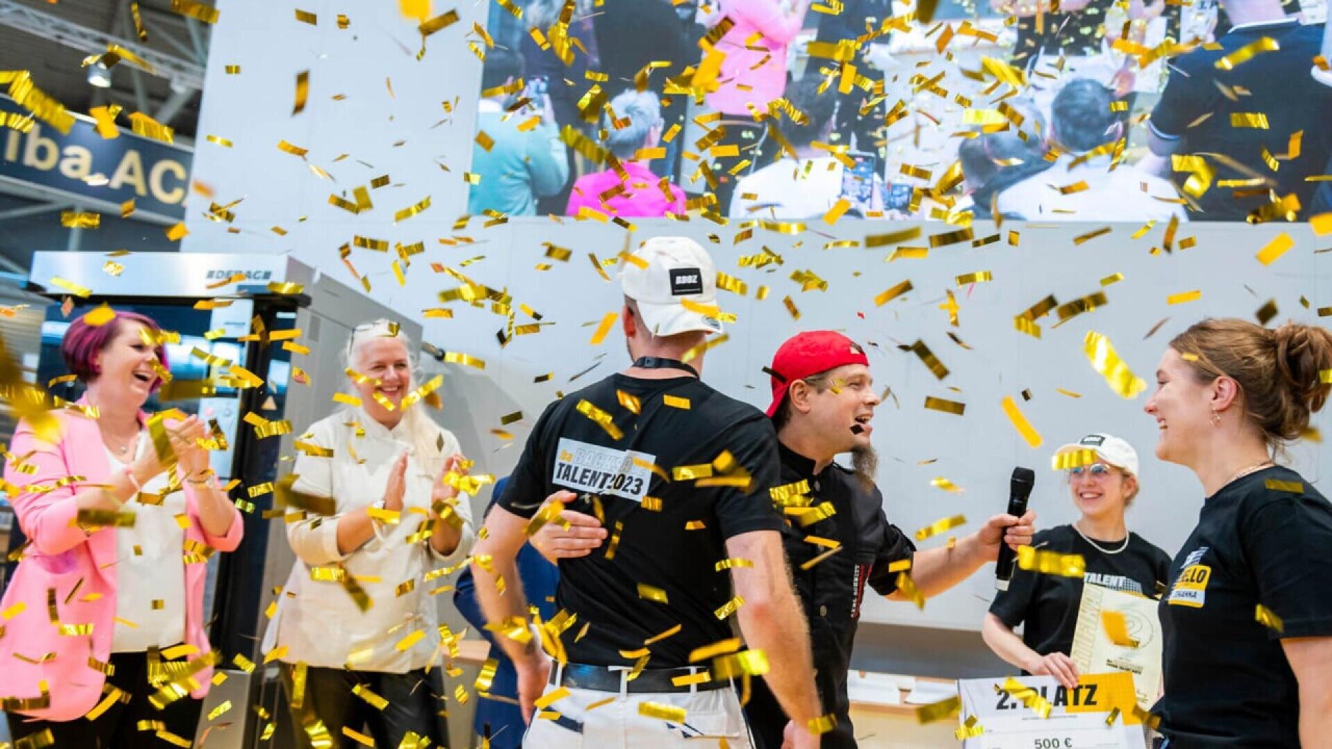 People celebrate on stage with confetti falling around them. Several individuals smile, clap, and hold a sign. A large screen in the background shows the same scene, enhancing the festive atmosphere.