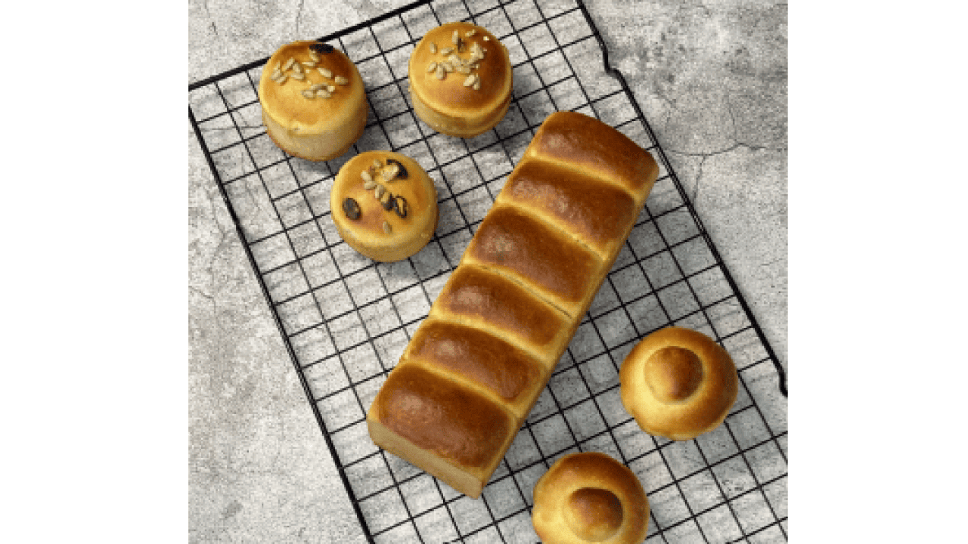 A golden-brown loaf of bread and six small round bread rolls, some topped with seeds, are cooling on a black wire rack set on a gray textured surface.