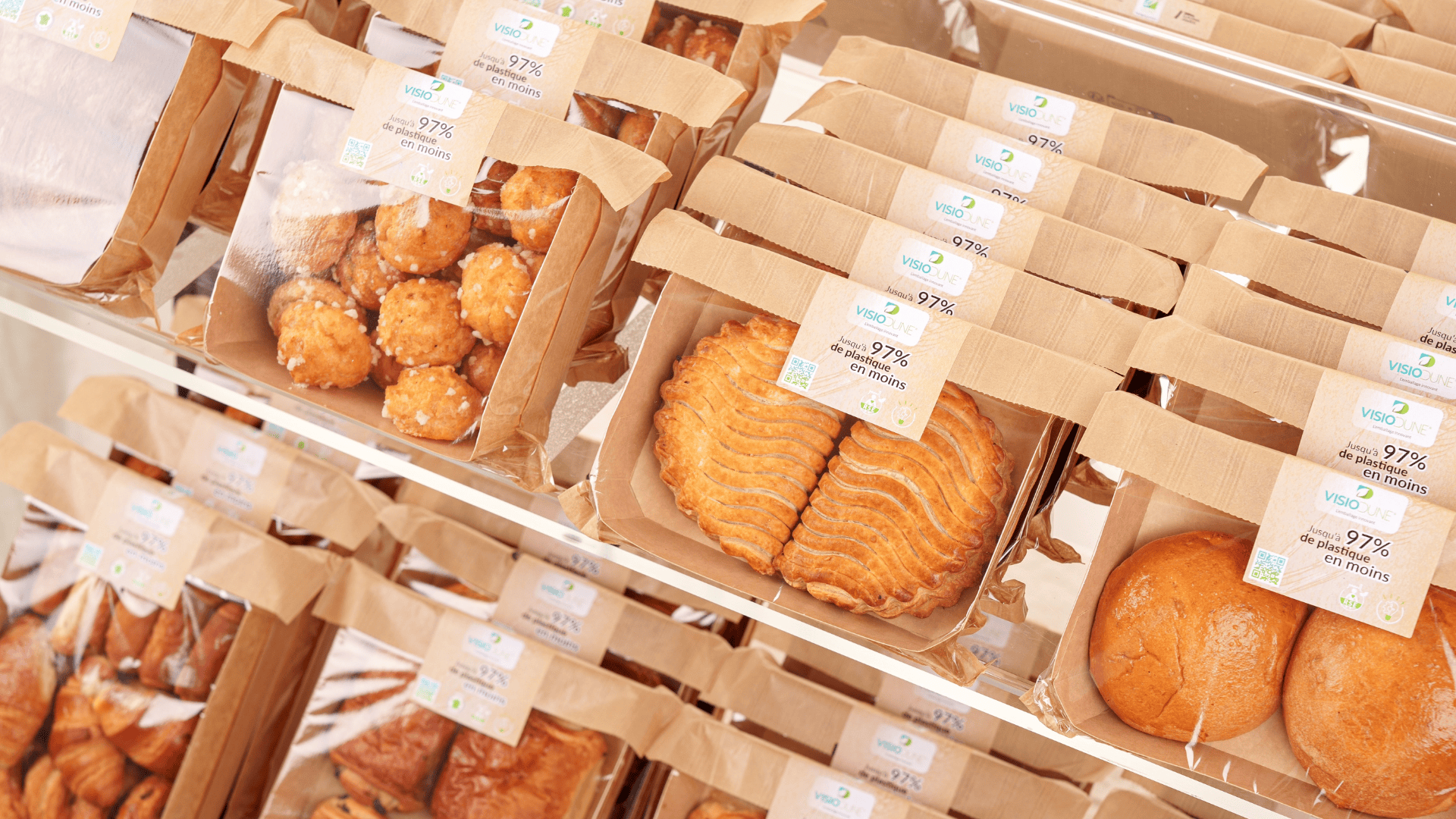 Assorted packaged pastries and baked goods are displayed on shelves, each in brown paper and plastic containers with white labels showing product details and prices.