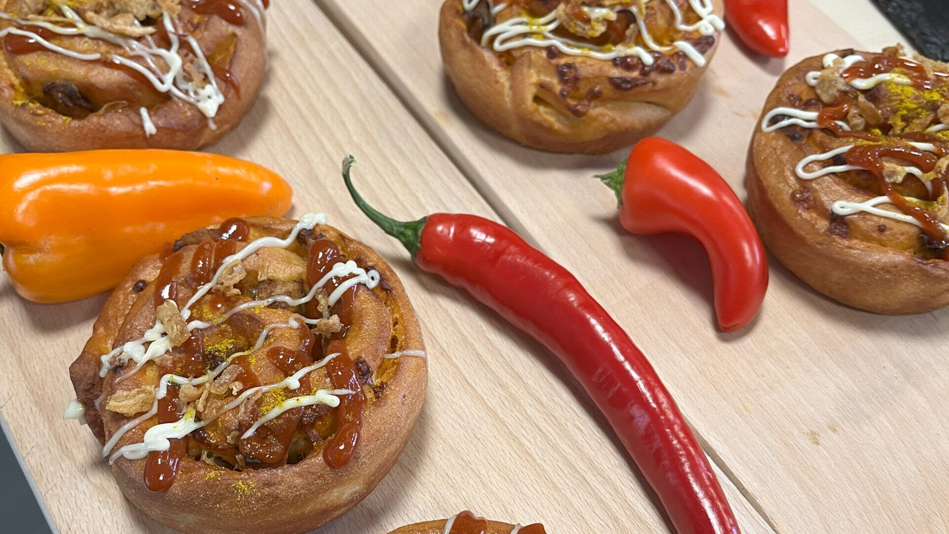 Drei herzhafte Teigrollen mit einem Spritzer Ketchup und Mayonnaise liegen auf einem Holzbrett, umgeben von einer roten Chilischote, einer roten Paprika und einer orangefarbenen Paprika.