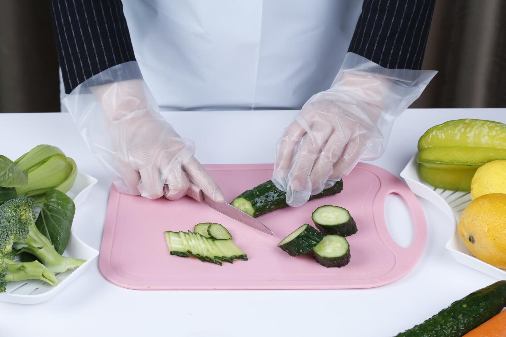 A person wearing clear gloves slices a cucumber on a pink cutting board. Plates of assorted vegetables and fruits, including broccoli, starfruit, and mango, are on the white table nearby.