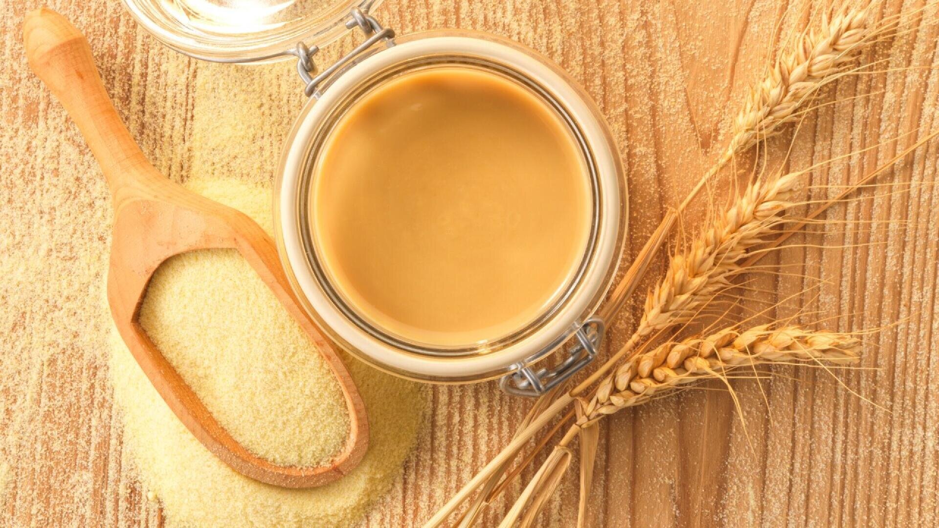 A glass jar of tahini sits on a wooden surface next to wheat stalks, a wooden scoop, and a small pile of semolina flour.