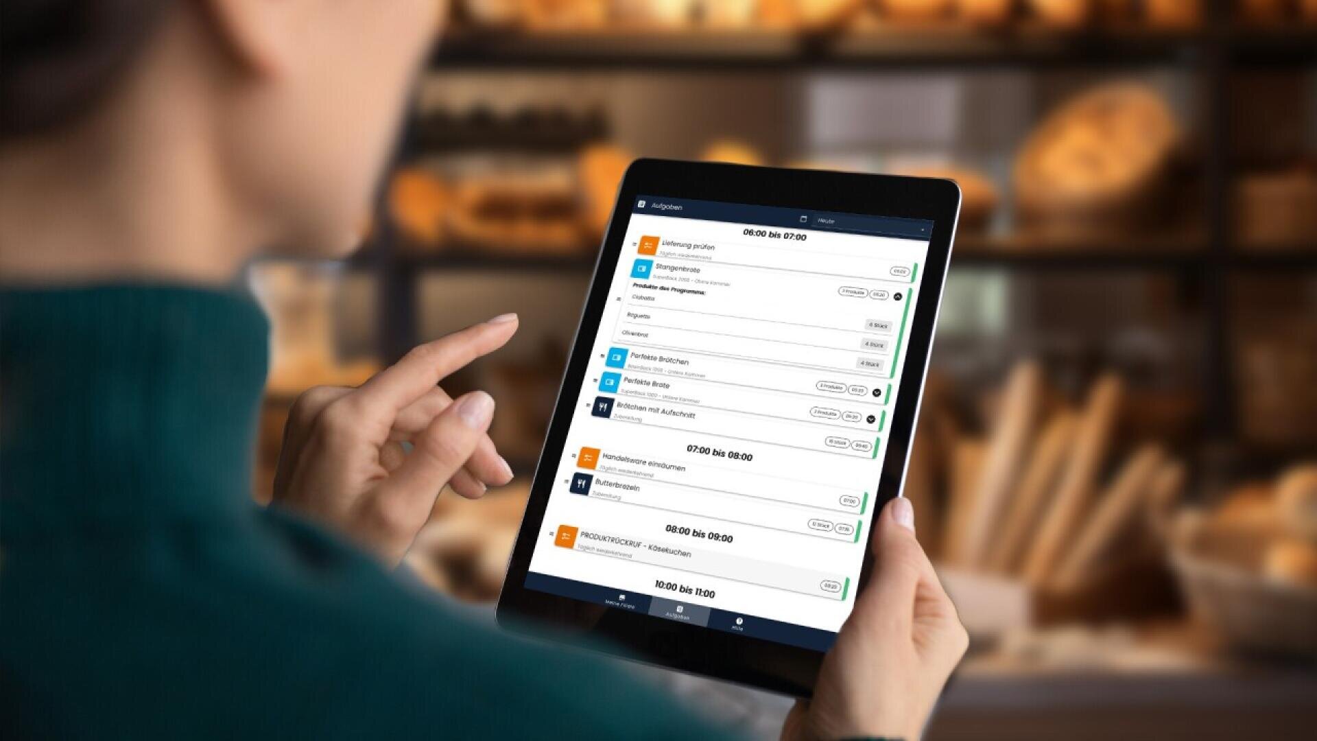 A person holds a tablet displaying a digital calendar or scheduling app, with various appointments listed. The background is blurred, showing shelves with bread and baked goods.