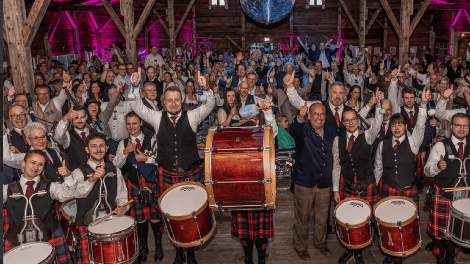 A large group of people cheers indoors as a drum band in plaid uniforms poses at the front, with wooden beams and pink lighting in the background.