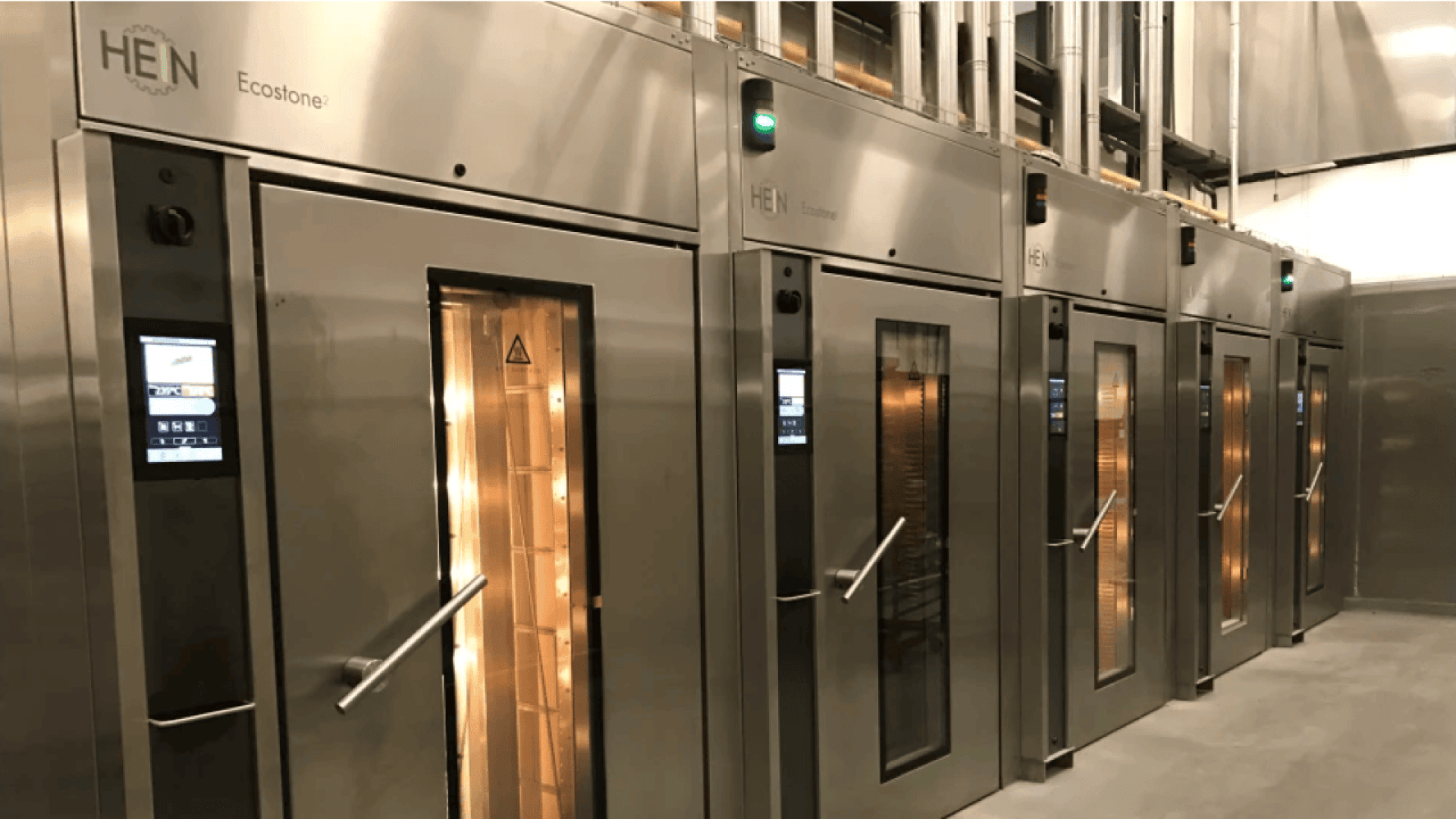 A row of large, stainless steel commercial ovens with glass doors and digital control panels lined up in a modern industrial kitchen. Green lights are illuminated above each oven door.