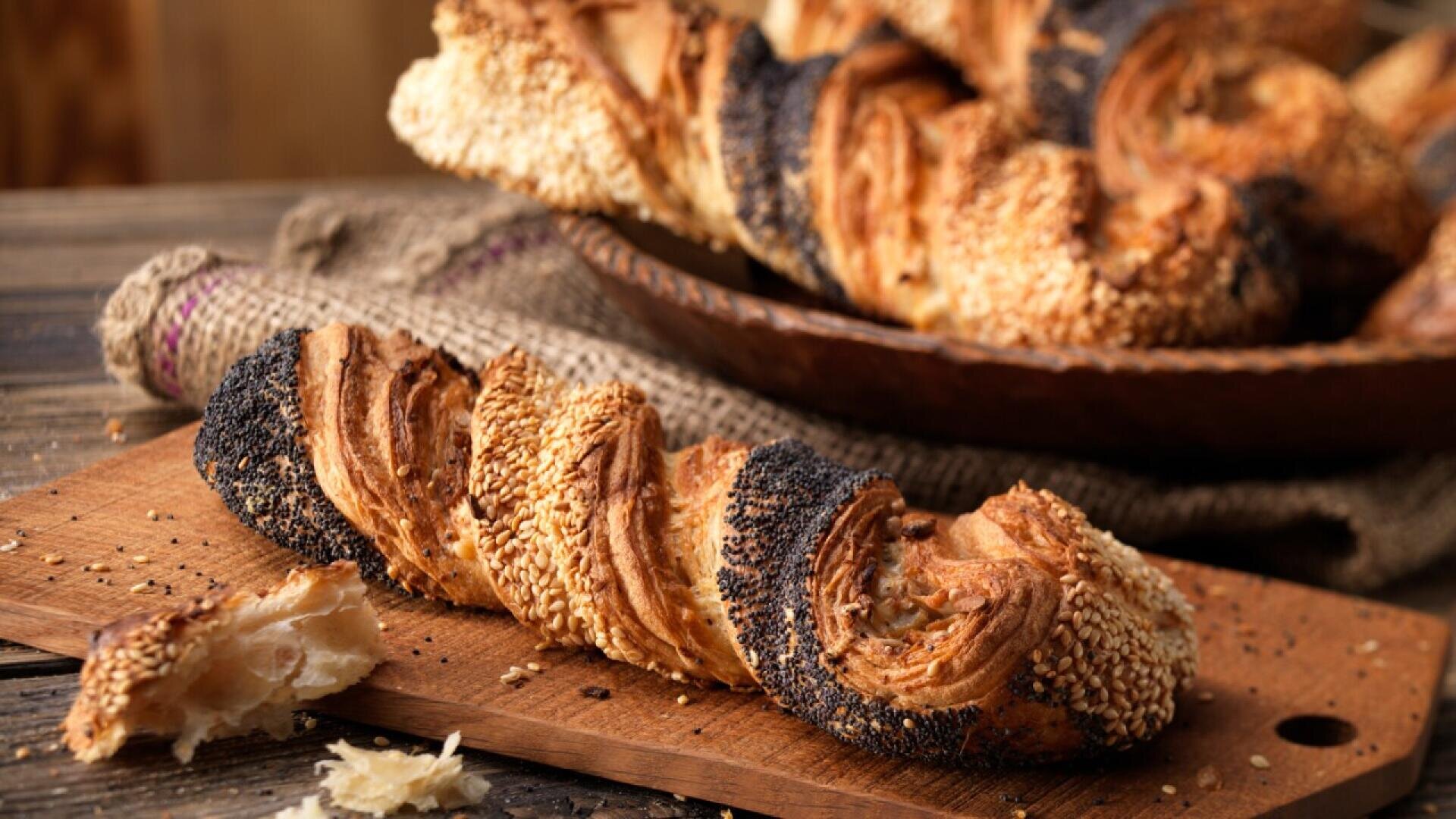A twisted, flaky bread loaf coated with sesame and poppy seeds sits on a wooden cutting board, with a few crumbs scattered nearby and more bread on a plate in the background.