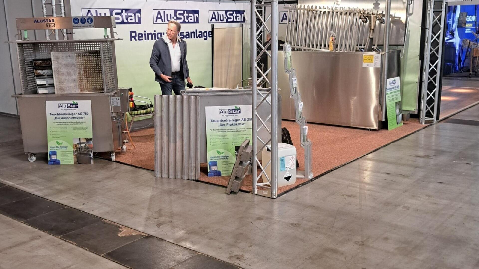 A man stands at an exhibition booth showcasing large industrial metal tanks and cleaning systems by AluStar. Informational signs and equipment components are displayed around the booth on a carpeted platform.
