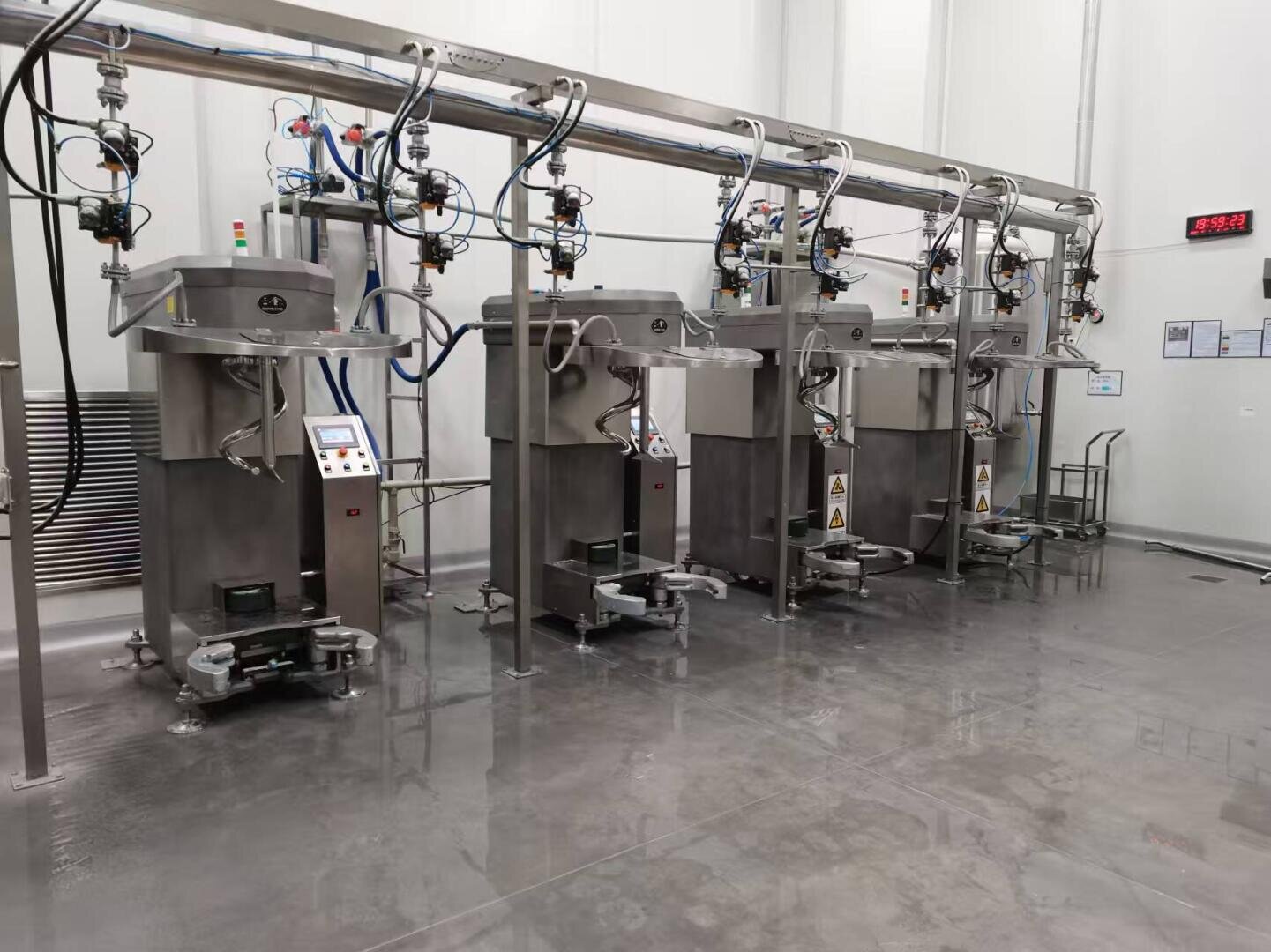 Four stainless steel industrial packaging machines are lined up in a clean, brightly lit facility. Overhead pipes and cables connect to each machine, and the floor is shiny and reflective. A digital clock is mounted on the wall.