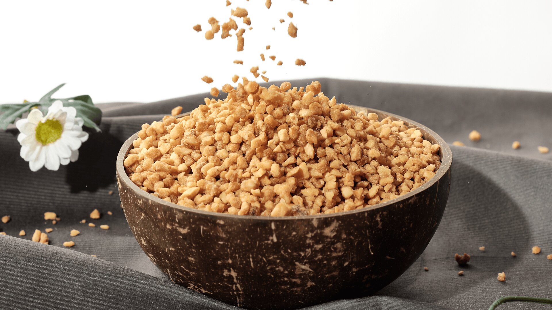 A coconut bowl filled with light brown granules, possibly chopped nuts or cereal, with more being poured in from above. A white flower decorates the background on a gray fabric surface.