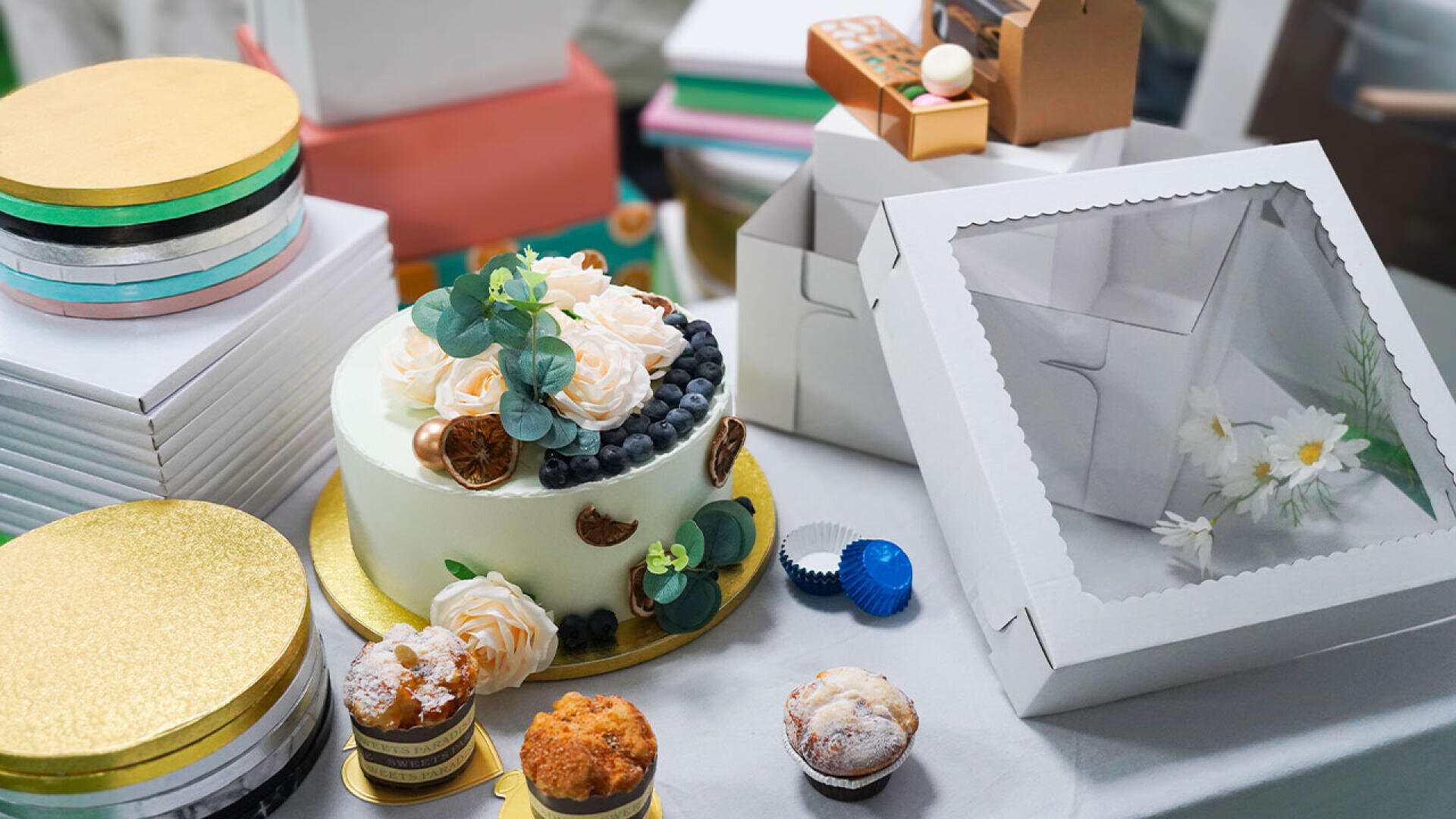 A decorated cake topped with flowers, blueberries, and gold accents sits on a table with muffins, empty pastry boxes, and stacked cake boards in the background.
