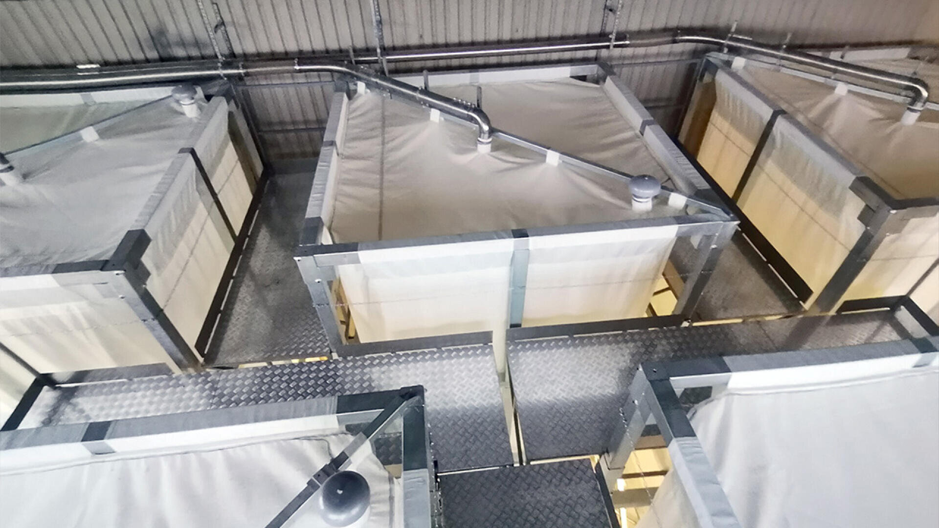 A top-down view of several large, rectangular industrial containers covered with white tarps, connected by metal pipes, and arranged on a metal platform inside a facility.