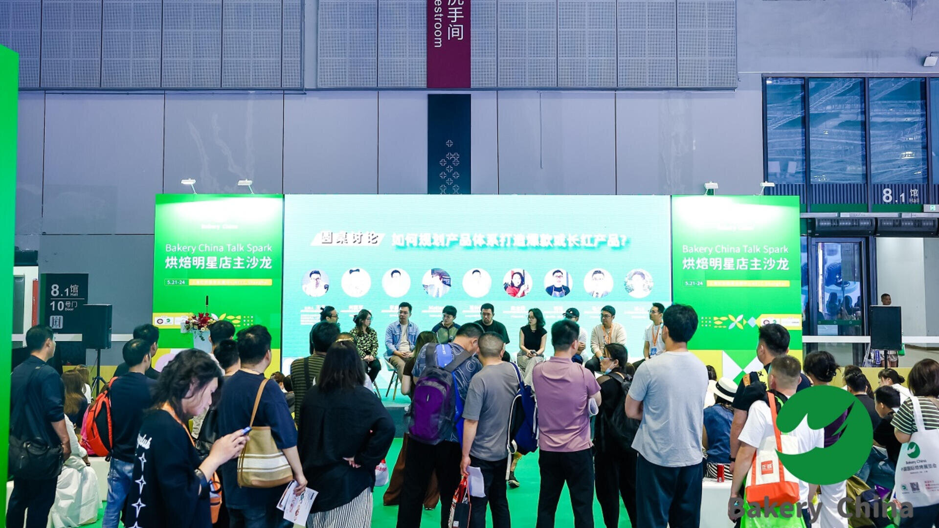 A group of people sit on a stage panel in front of a green backdrop with Chinese and English text, while an audience stands and watches at an indoor event or conference.
